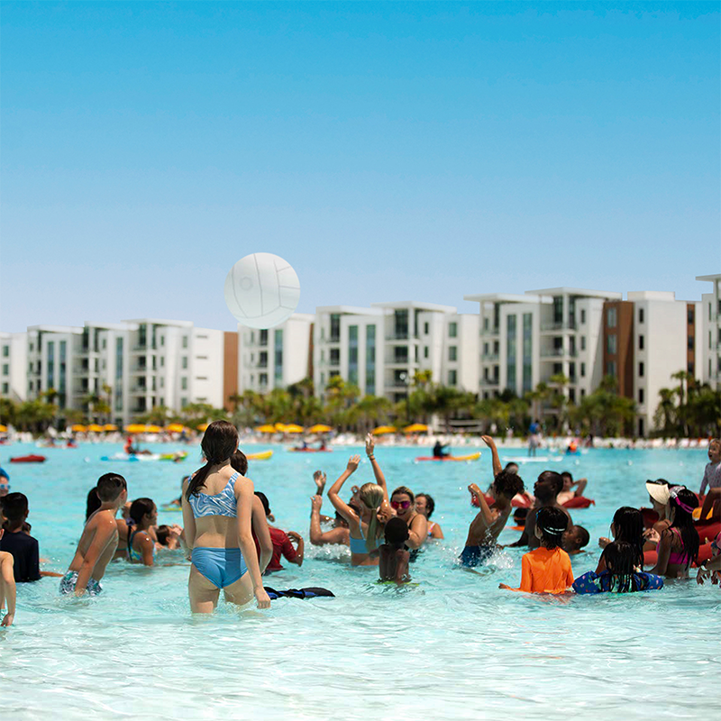 Children in swimwear play with a volleyball in a large pool with buildings in the background.