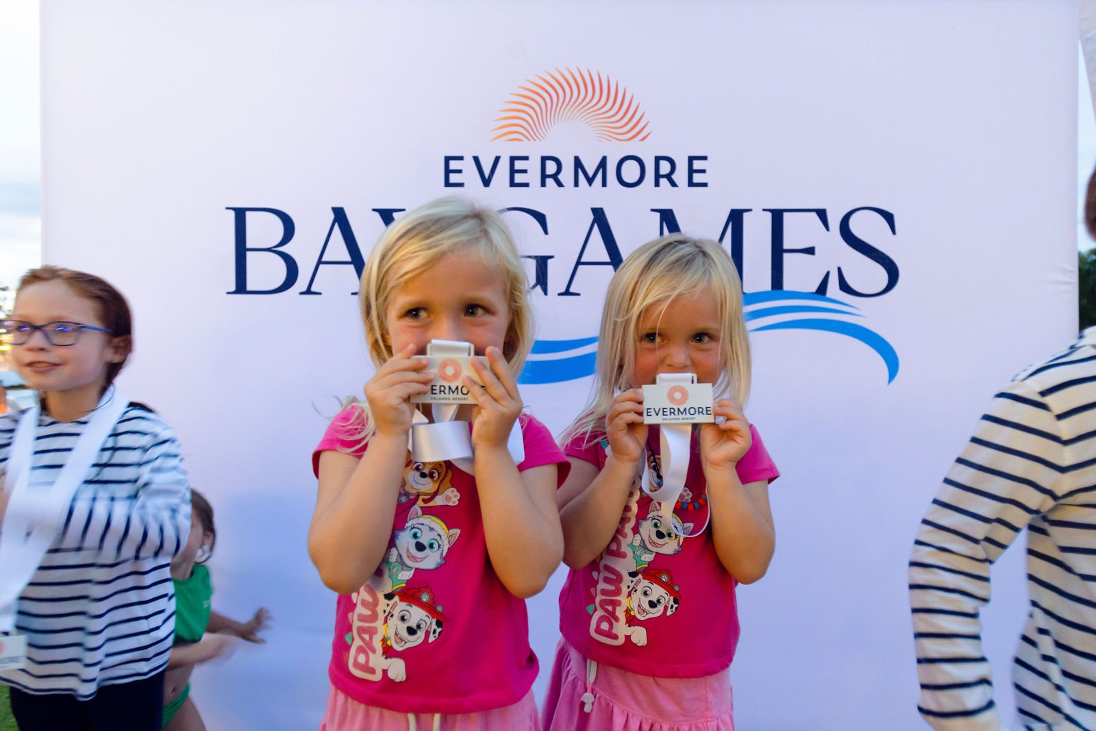 Two young girls in pink Paw Patrol shirts holding Evermore Bay Games medals in front of event banner.