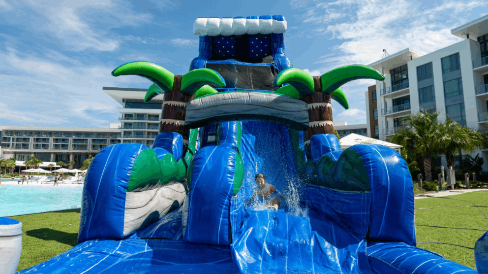 Child sliding down a blue inflatable water slide with palm tree decorations at a resort.