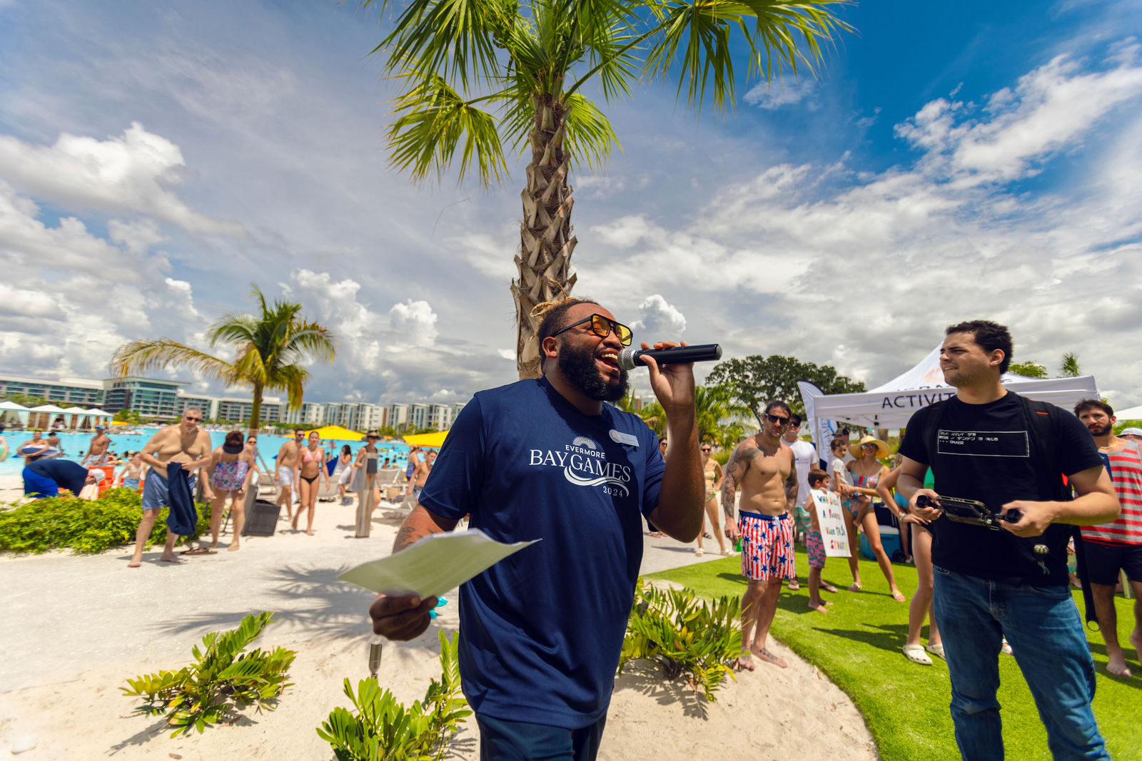 Man speaking into microphone at sunny outdoor pool event with people and palm trees around him.