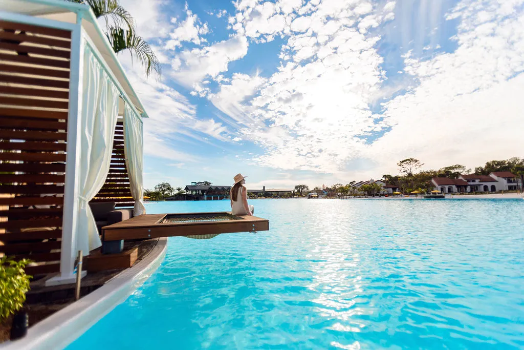 Woman in white dress and hat sitting on a wooden platform over a large swimming pool under blue sky.