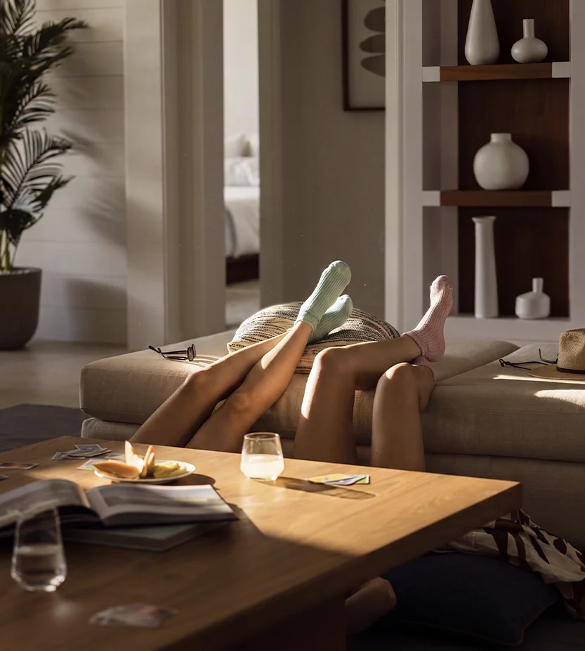 Two people lying on floor with legs raised on sofa, wearing pastel-colored socks in sunlit living room.