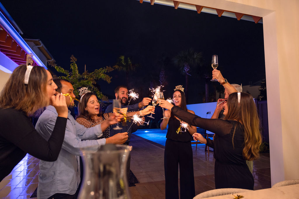 Group of friends celebrating at night with sparklers and champagne near a lit swimming pool.