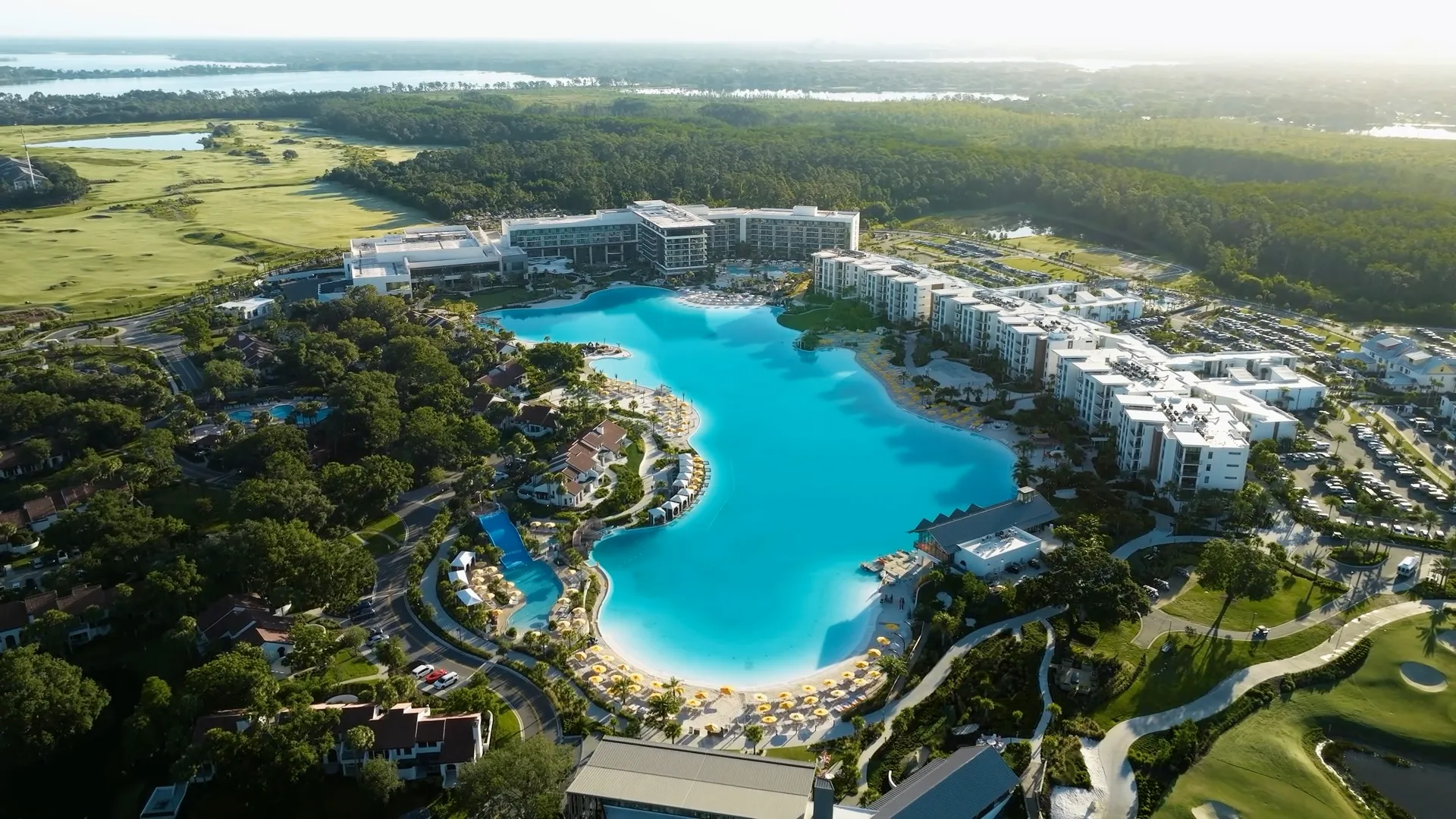 Aerial view of a large resort around a bright blue lagoon pool with surrounding buildings and greenery.