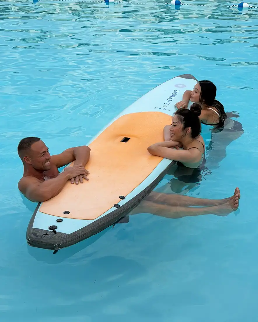 Three people in a pool leaning on an orange and white paddleboard, smiling and relaxing together.