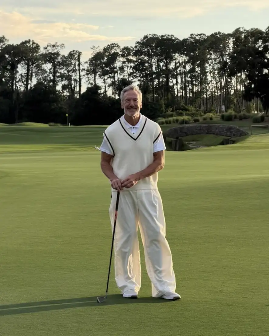 Older man in white golf attire standing on green grass holding a golf club with trees and bridge behind.