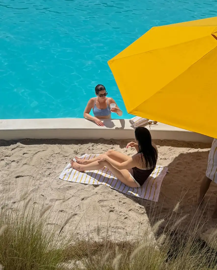 Woman in pool holding a drink while another sits on sand under a large yellow umbrella.