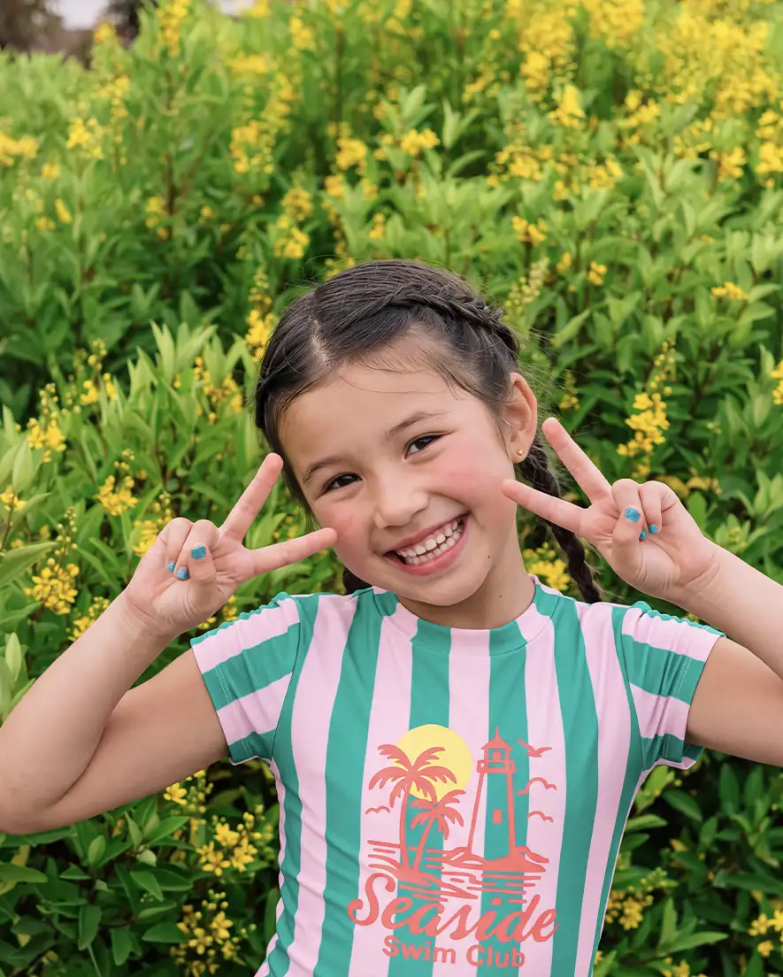 Smiling girl with braided hair making peace signs with both hands in front of green plants with yellow flowers.