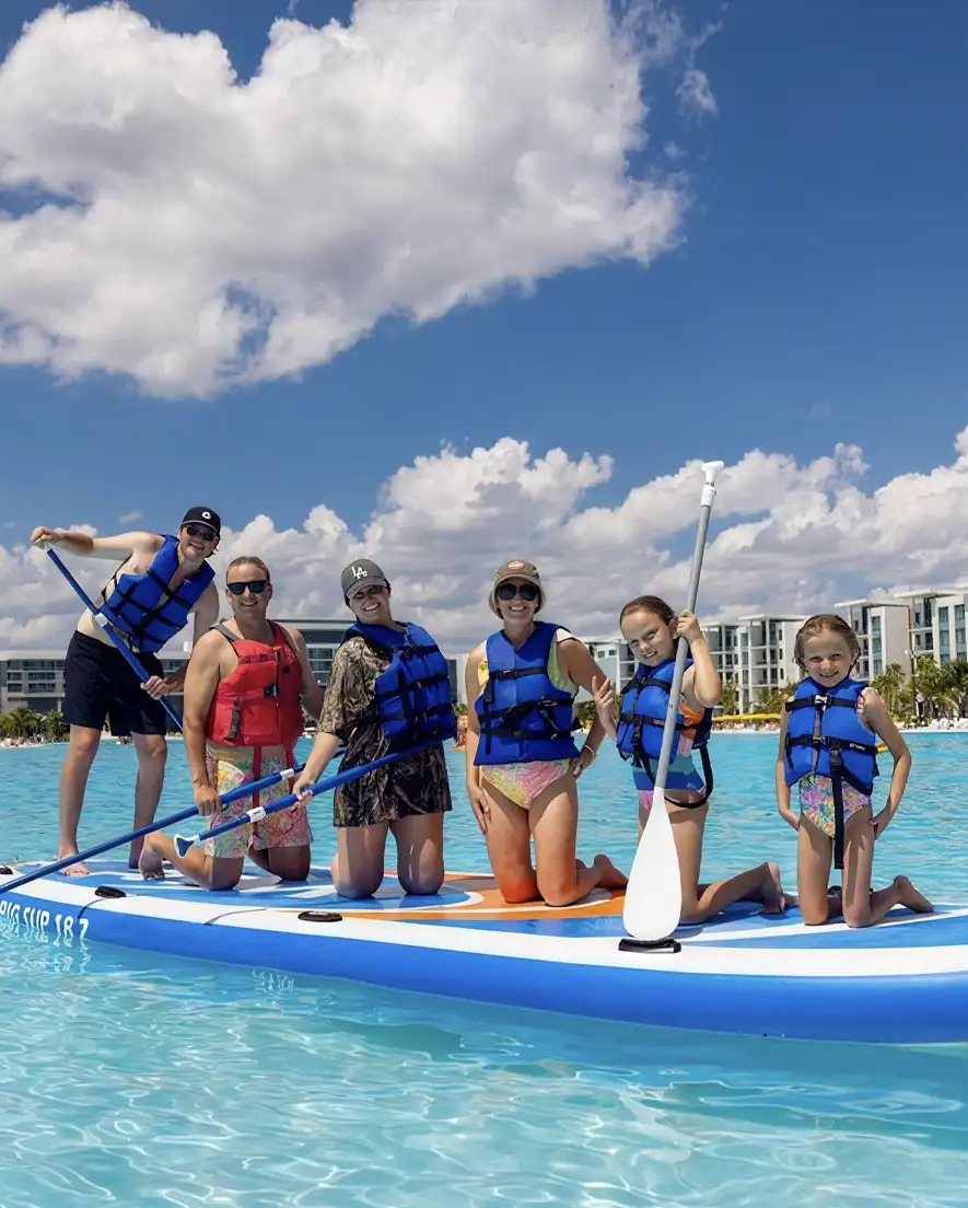 Six people wearing life jackets kneeling on a large paddleboard in clear blue water near buildings.