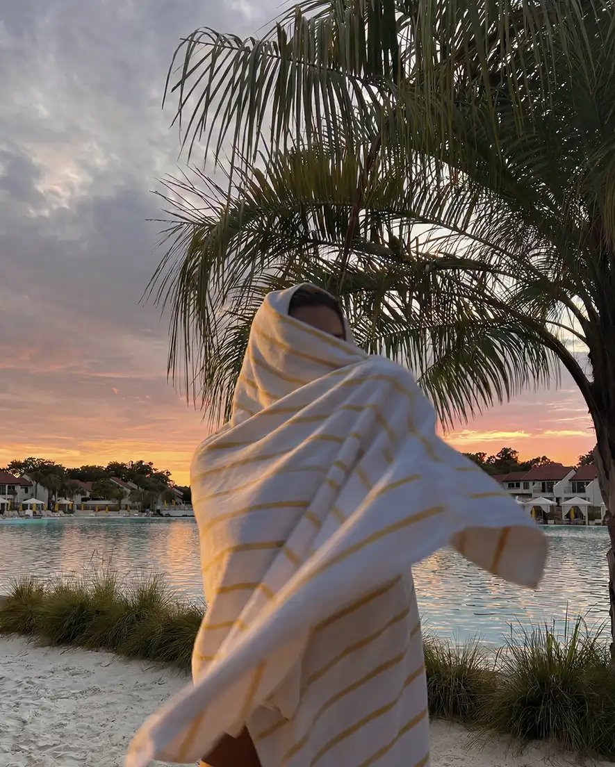 Person wrapped in striped towel near a pool with palm trees at sunset.