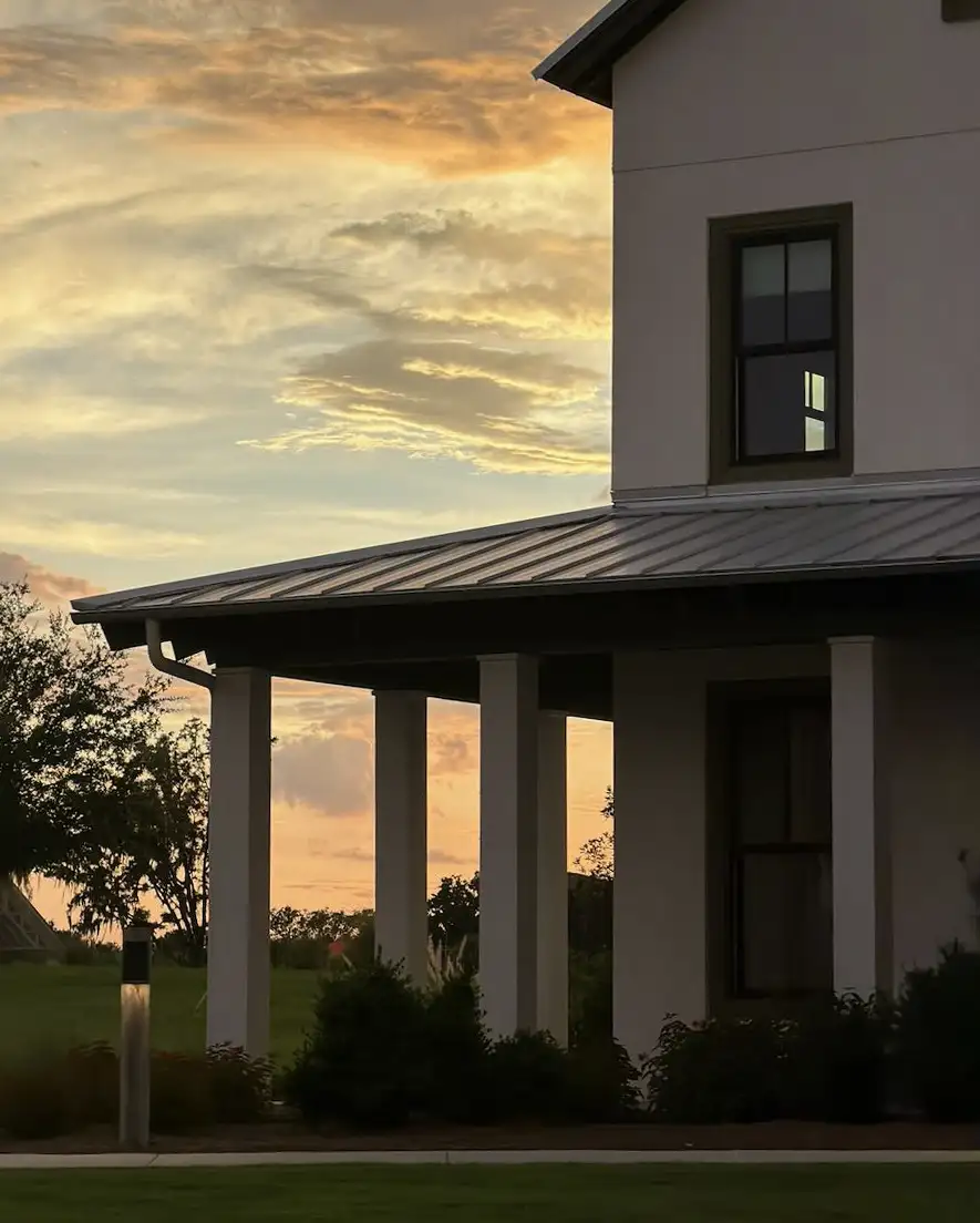 Part of a house with white pillars and a metal roof at sunset with a partly cloudy sky.
