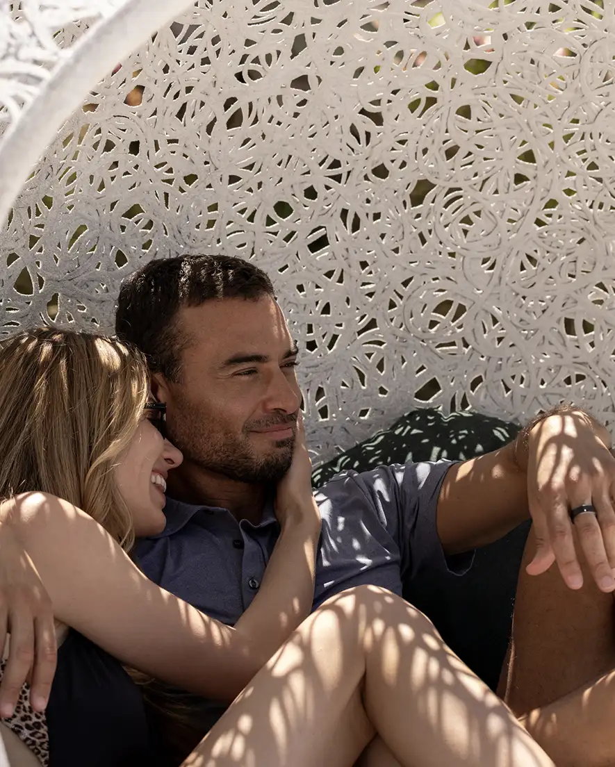 Couple sitting closely in an intricately patterned white outdoor chair with sunlight casting shadows.