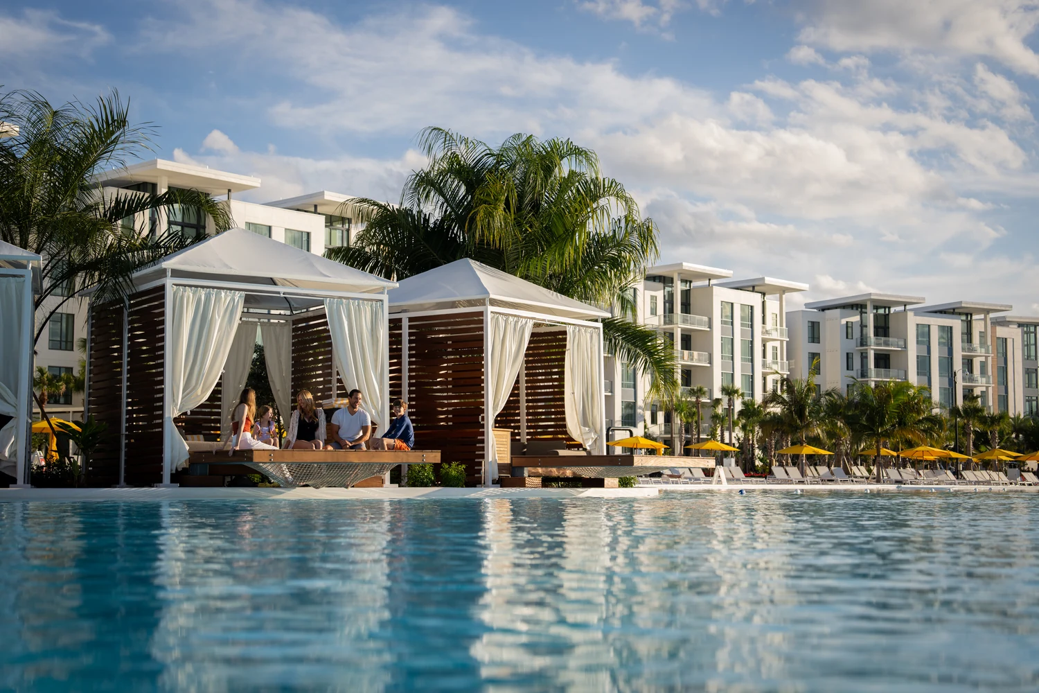 Family sitting and chatting in a white cabana by a pool at a modern resort with palm trees and buildings.