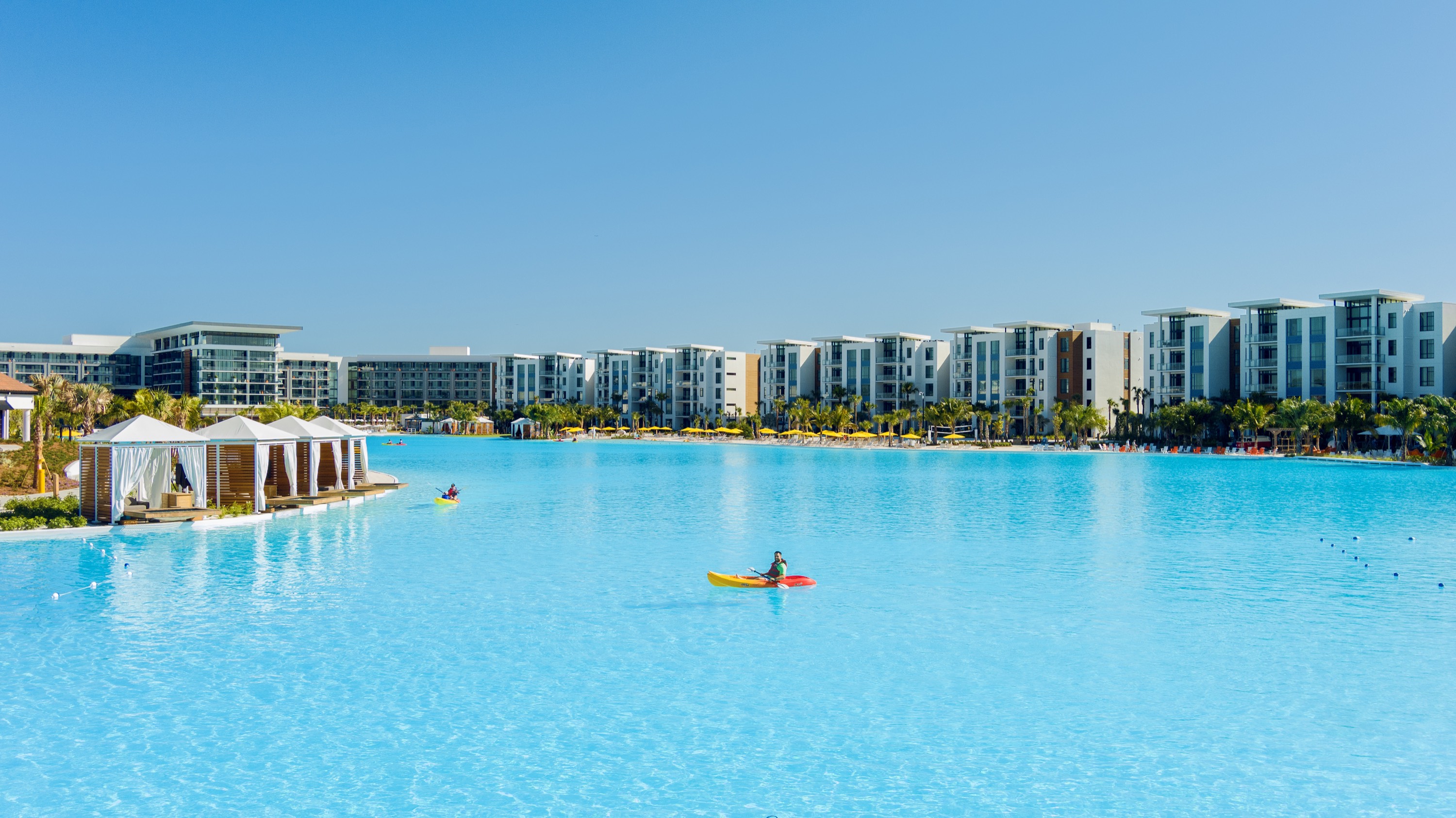 Person kayaking on a large pool with resort buildings and cabanas under clear blue sky.
