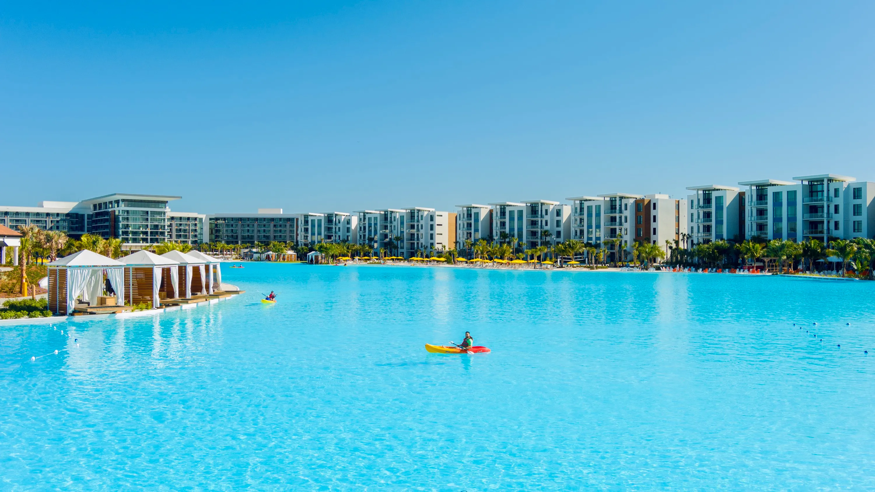 Person kayaking on a large pool with resort buildings and cabanas under clear blue sky.