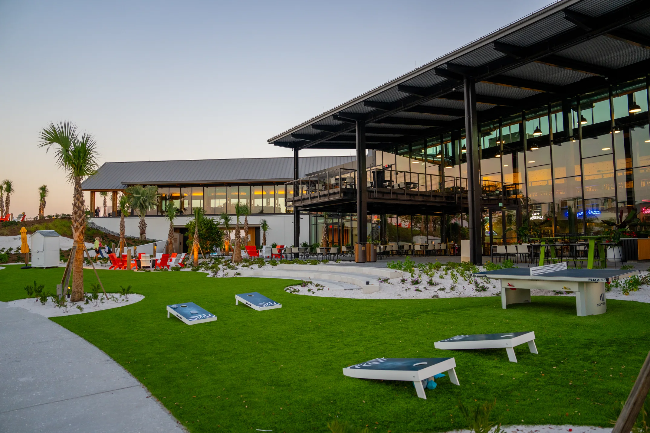 Outdoor lawn area with bean bag toss boards, ping pong table, palm trees, and a modern glass building.
