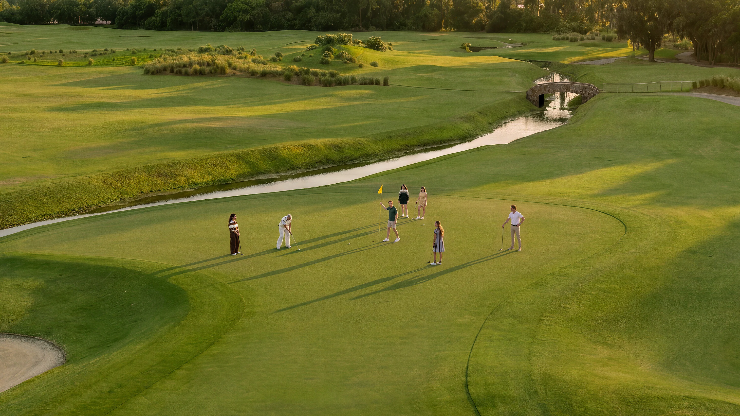 Six people playing golf on a green near a small stone bridge and creek at sunset.