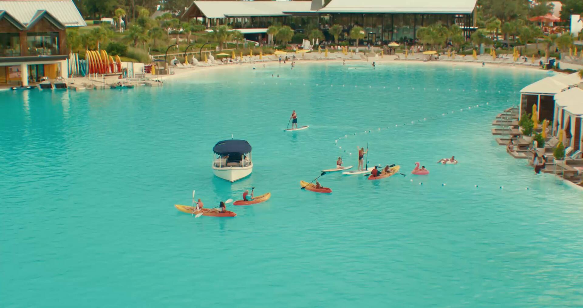 People kayaking, paddleboarding, and floating on a large turquoise lagoon near cabanas and buildings.