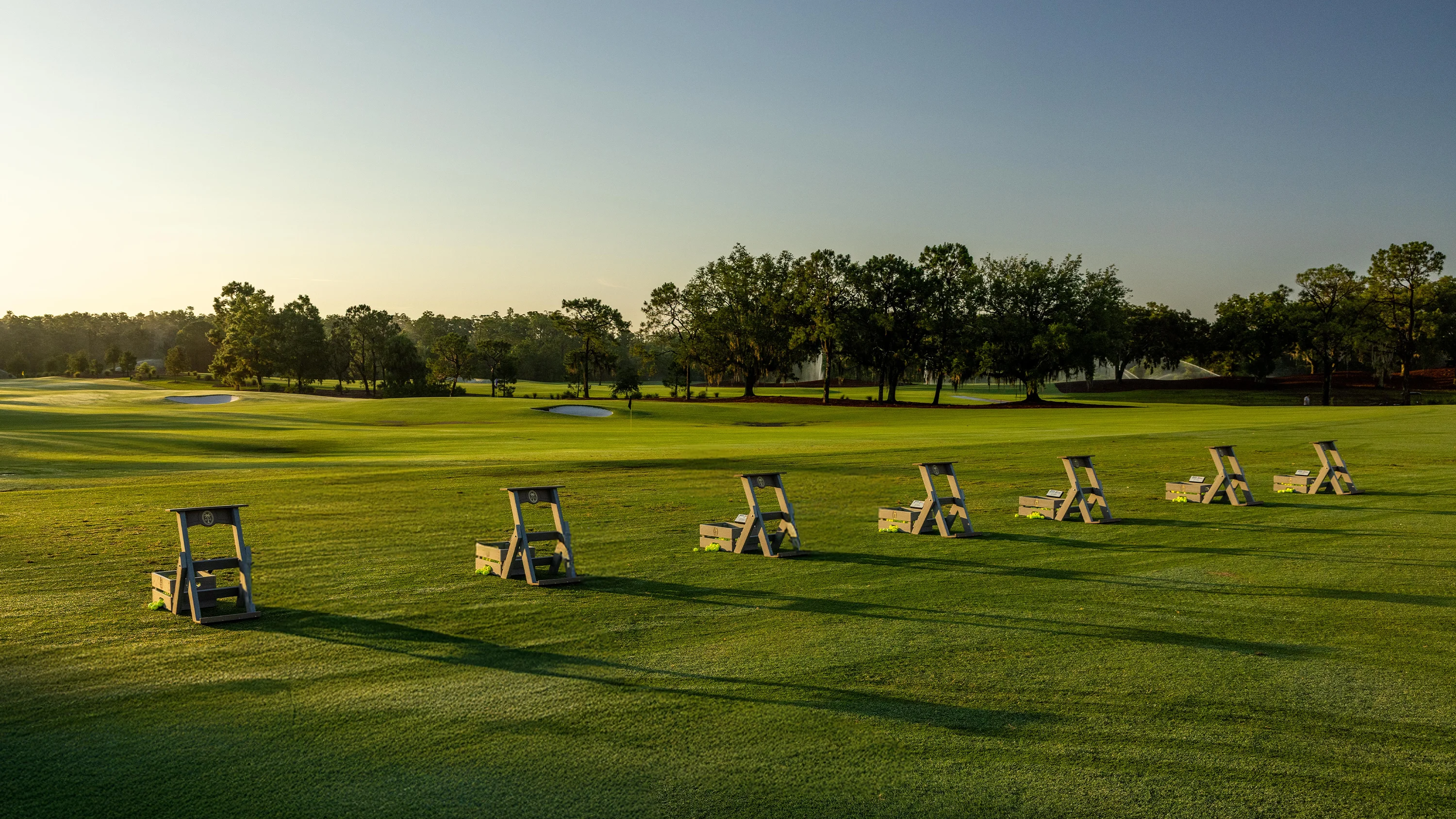 Row of wooden golf ball holders with balls aligned on a green driving range in early morning light.
