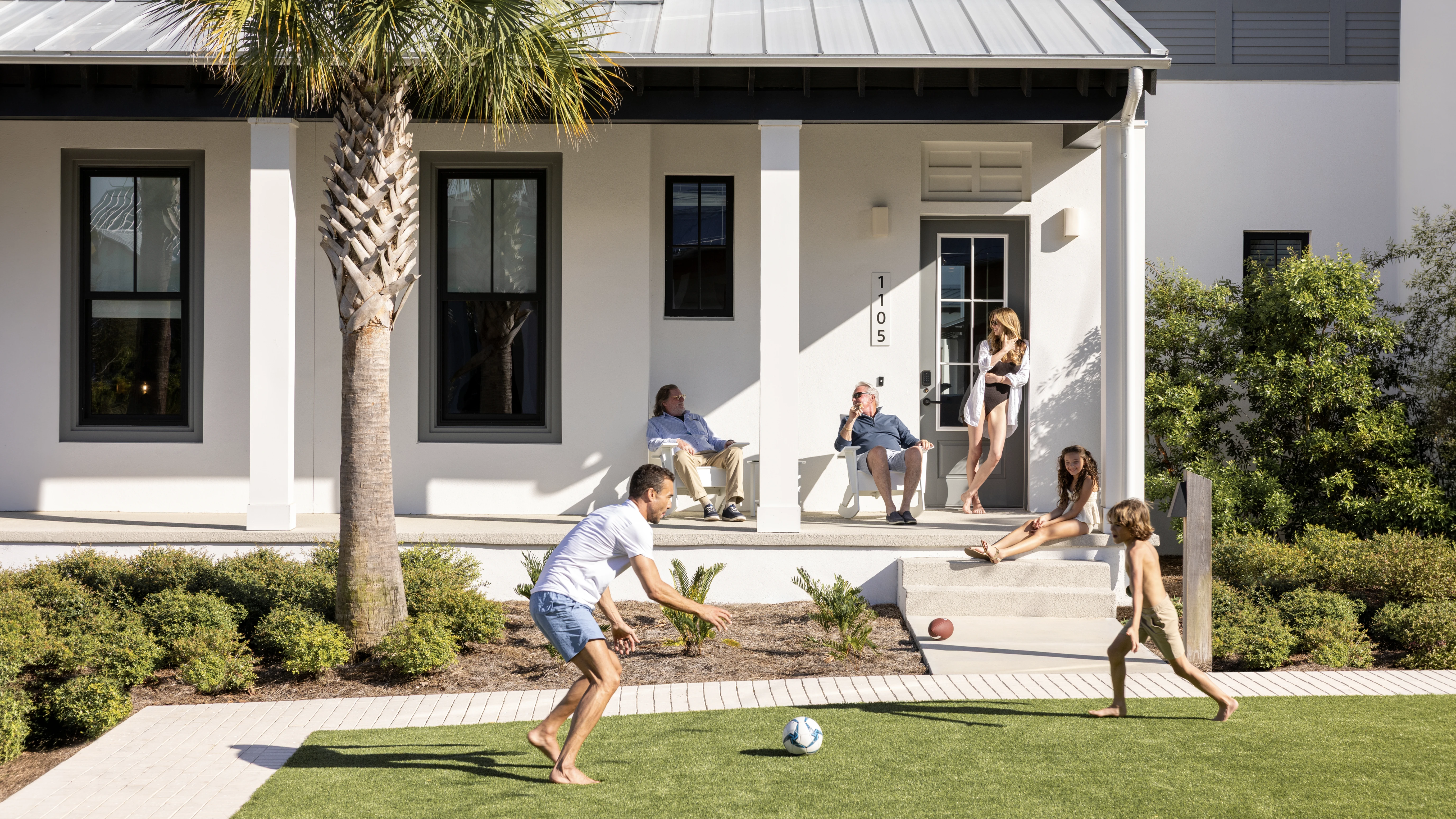 Family playing soccer on lawn outside an Evermore house with a porch and palm tree in yard.
