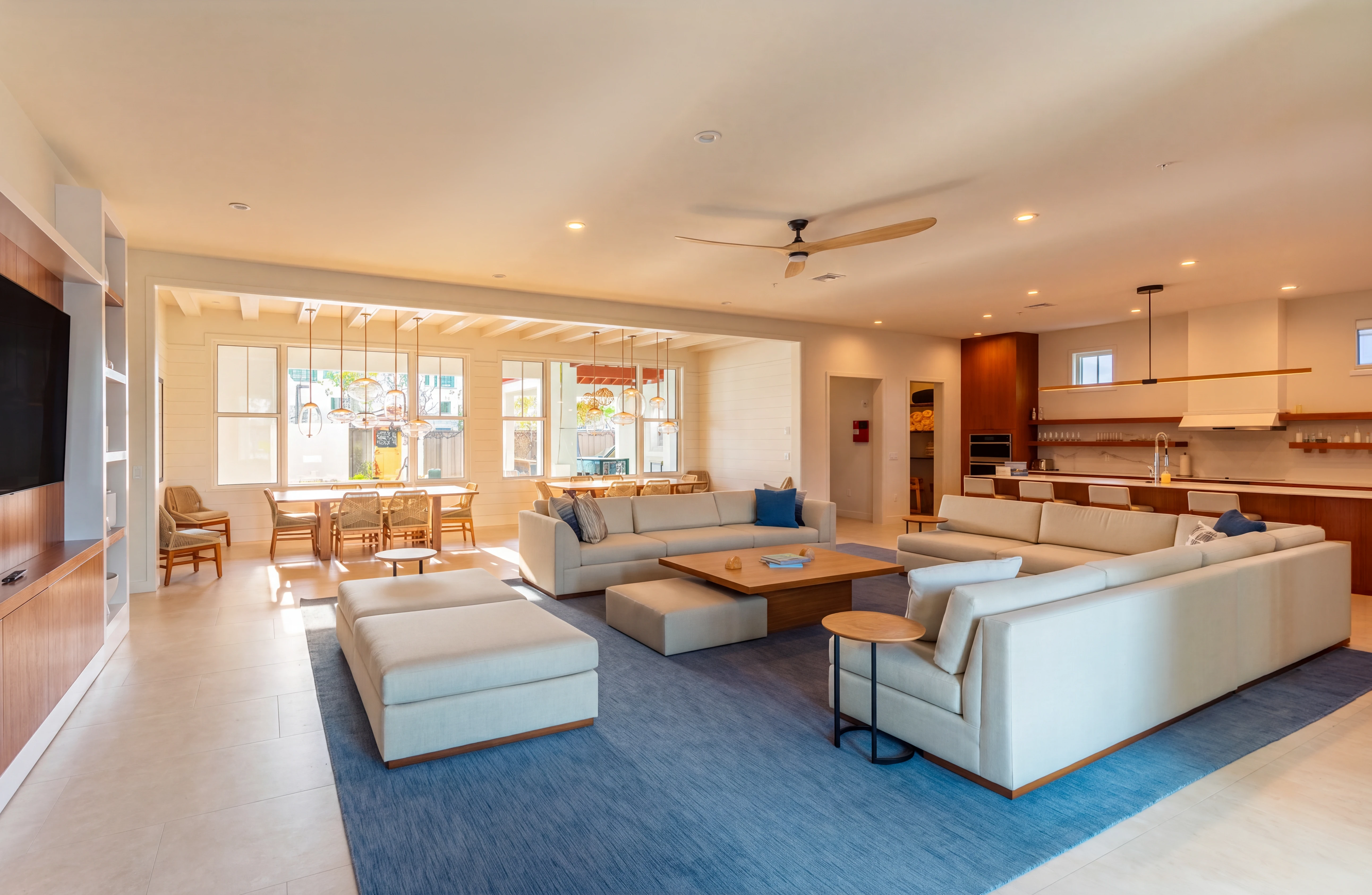 Spacious modern living room with beige sofas, wooden tables, blue rug, dining area, and kitchen in background.