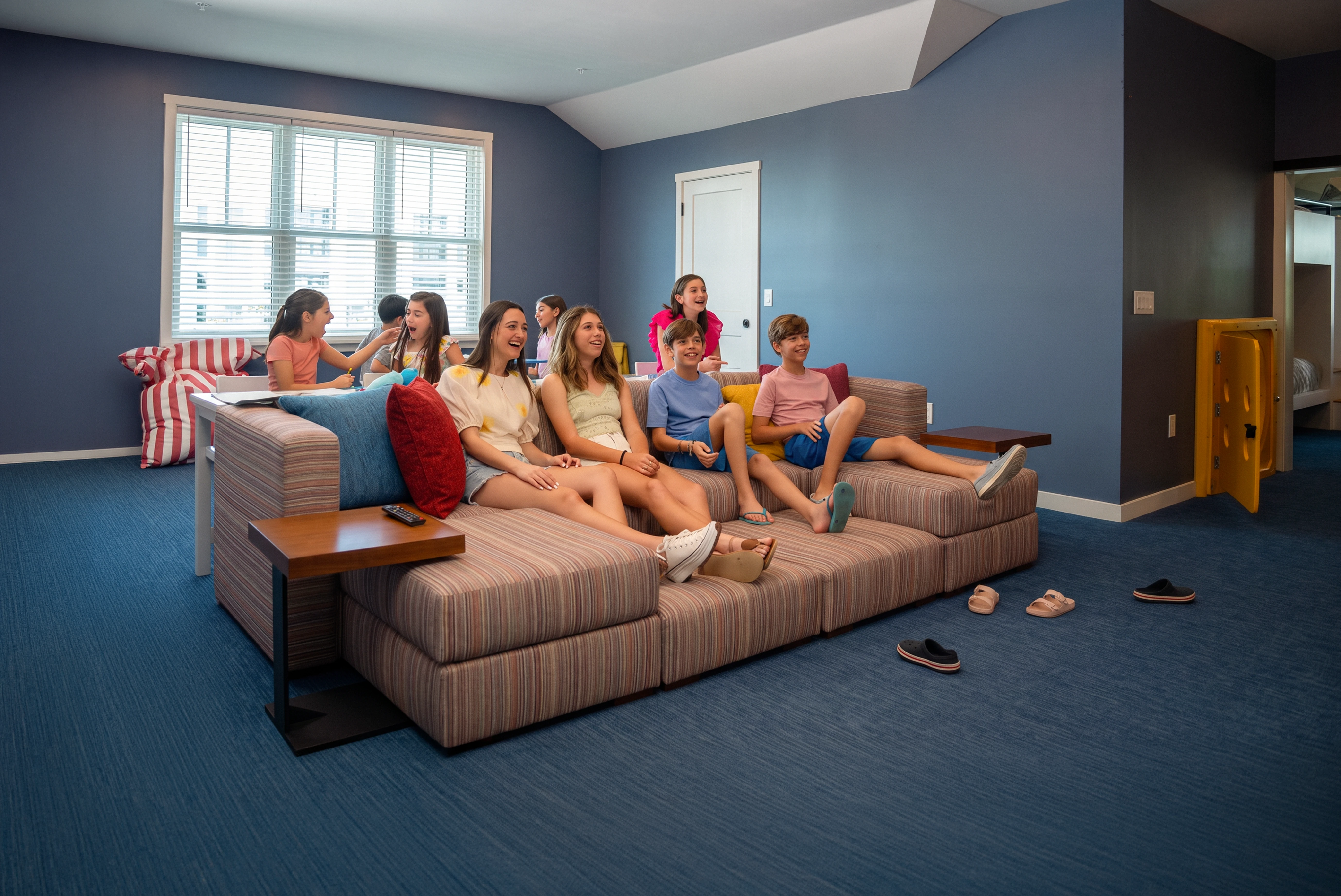 Seven kids sitting on couch and at table in a blue carpeted room with striped couch and seating.