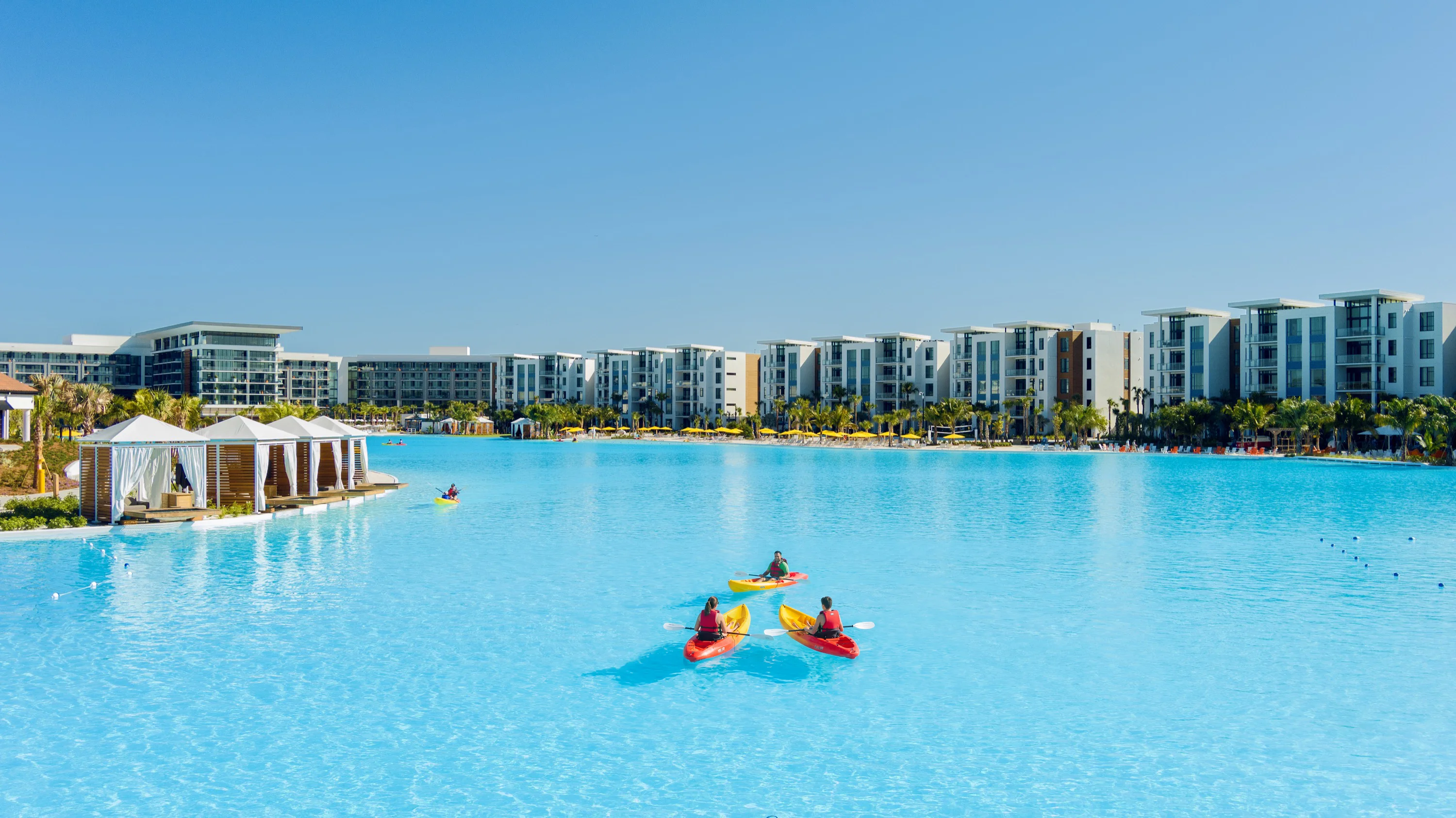 Three kayakers paddling on a large clear blue water lagoon with modern buildings and cabanas in background.