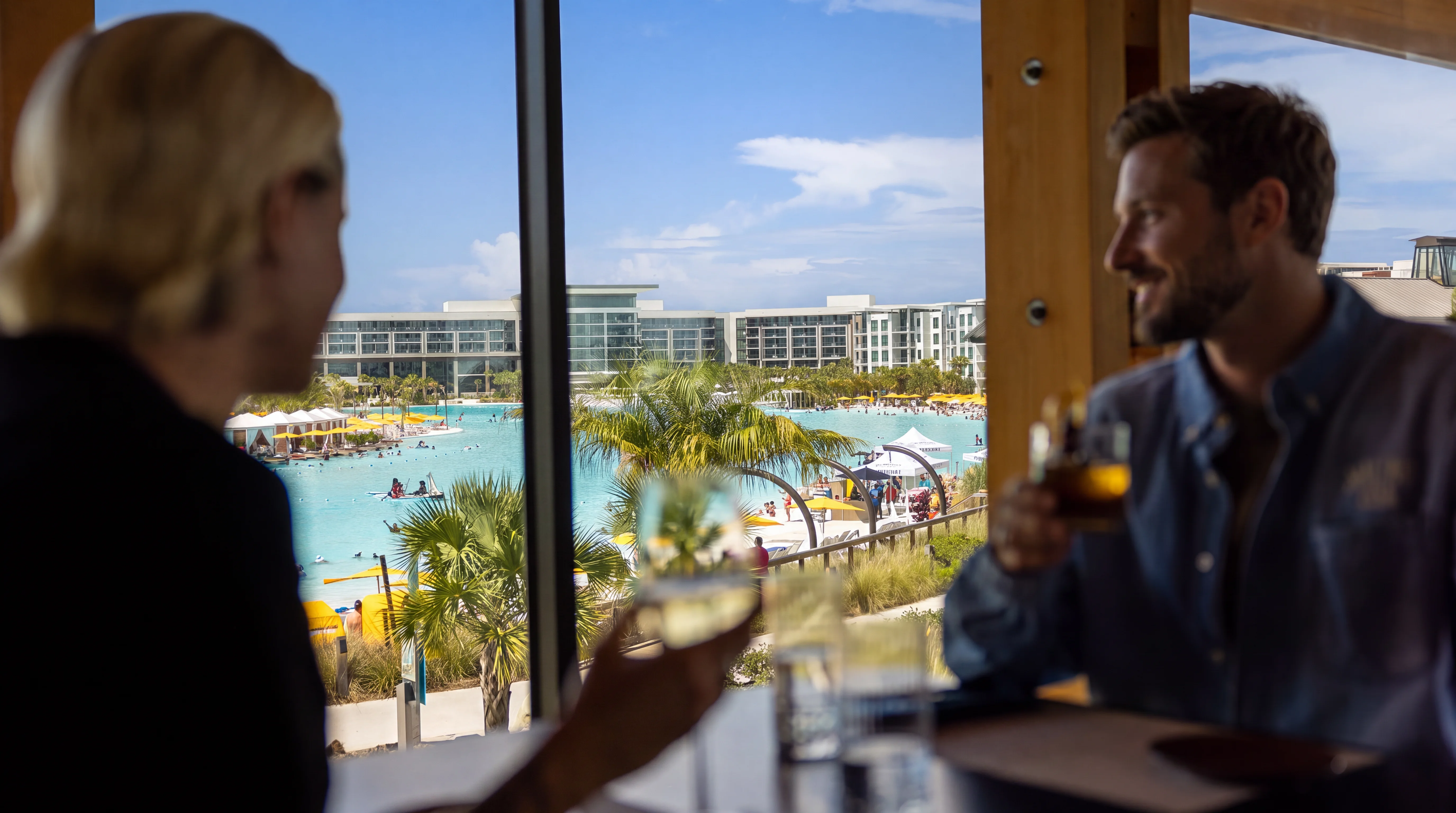 Two people inside a restaurant (Twin Views at Evermore) enjoying drinks with a bay and resort buildings outside the window.
