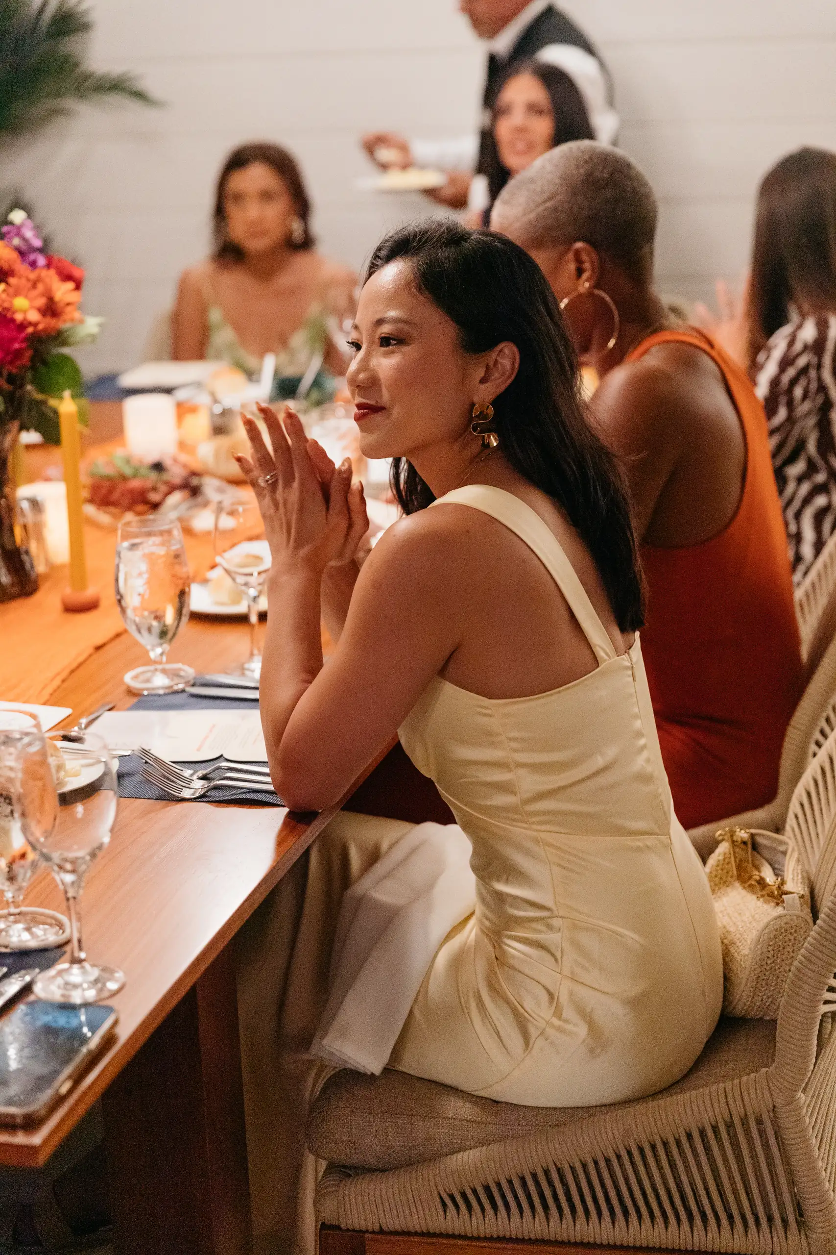 Woman in a pale yellow dress sitting at a dinner table with others, hands clasped, smiling contemplatively.
