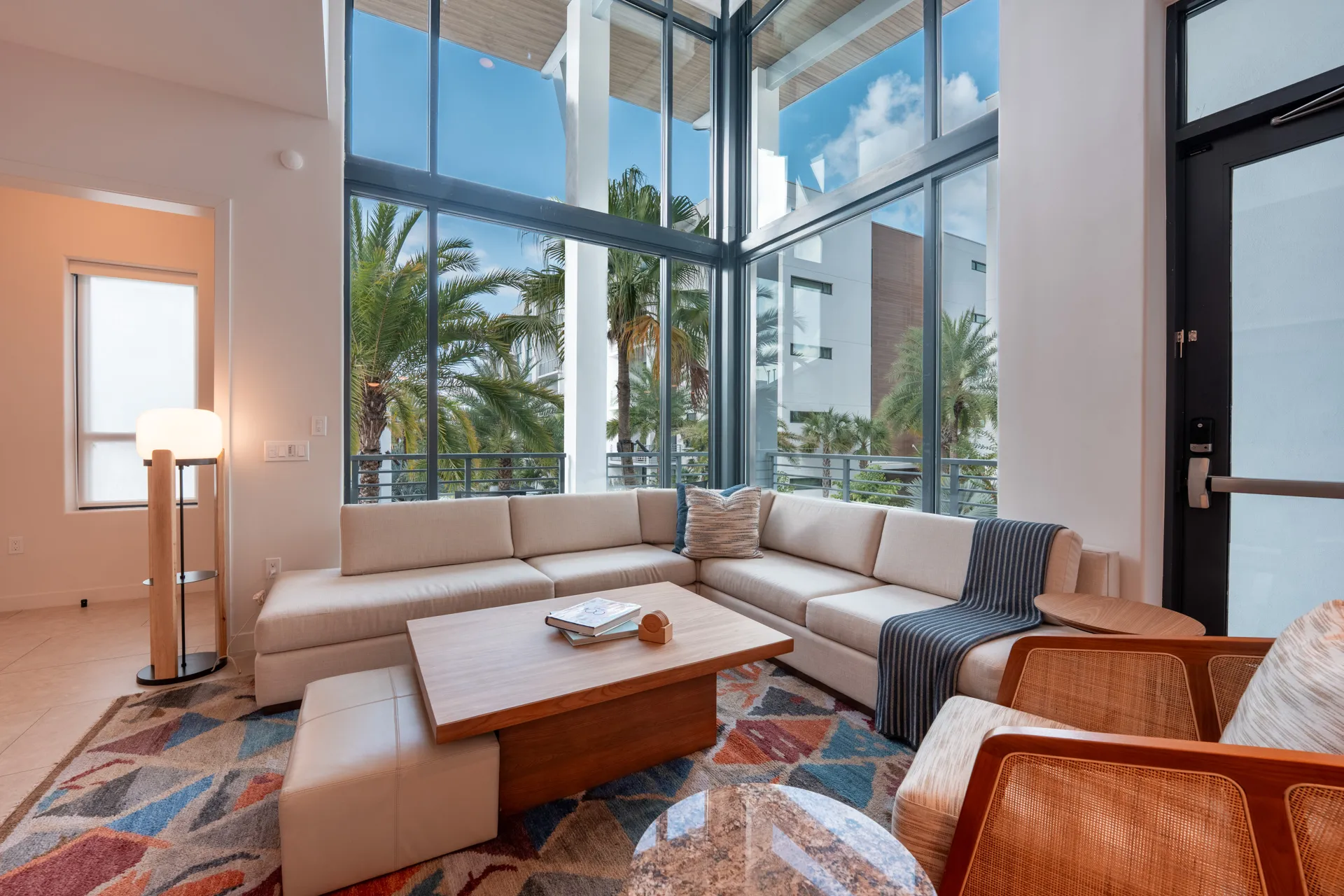 Modern living room with beige sectional sofa, wooden coffee table, patterned rug, and large windows with outdoor palm view.