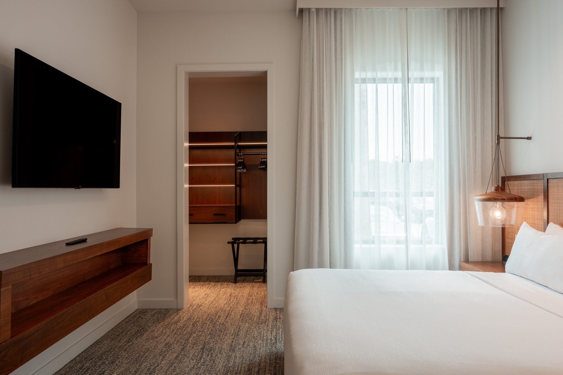 Modern bedroom with white bed, hanging lamp, wall-mounted TV, and open walk-in closet with wooden shelves.