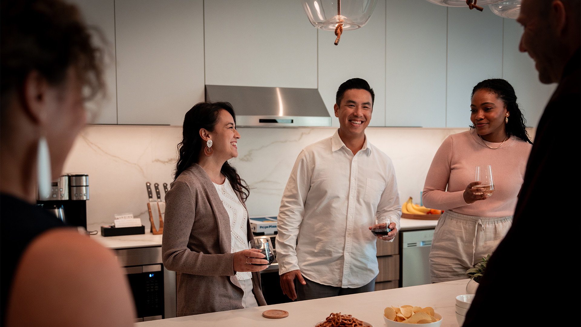Four people standing in a kitchen chatting and holding drinks with snacks on the counter.