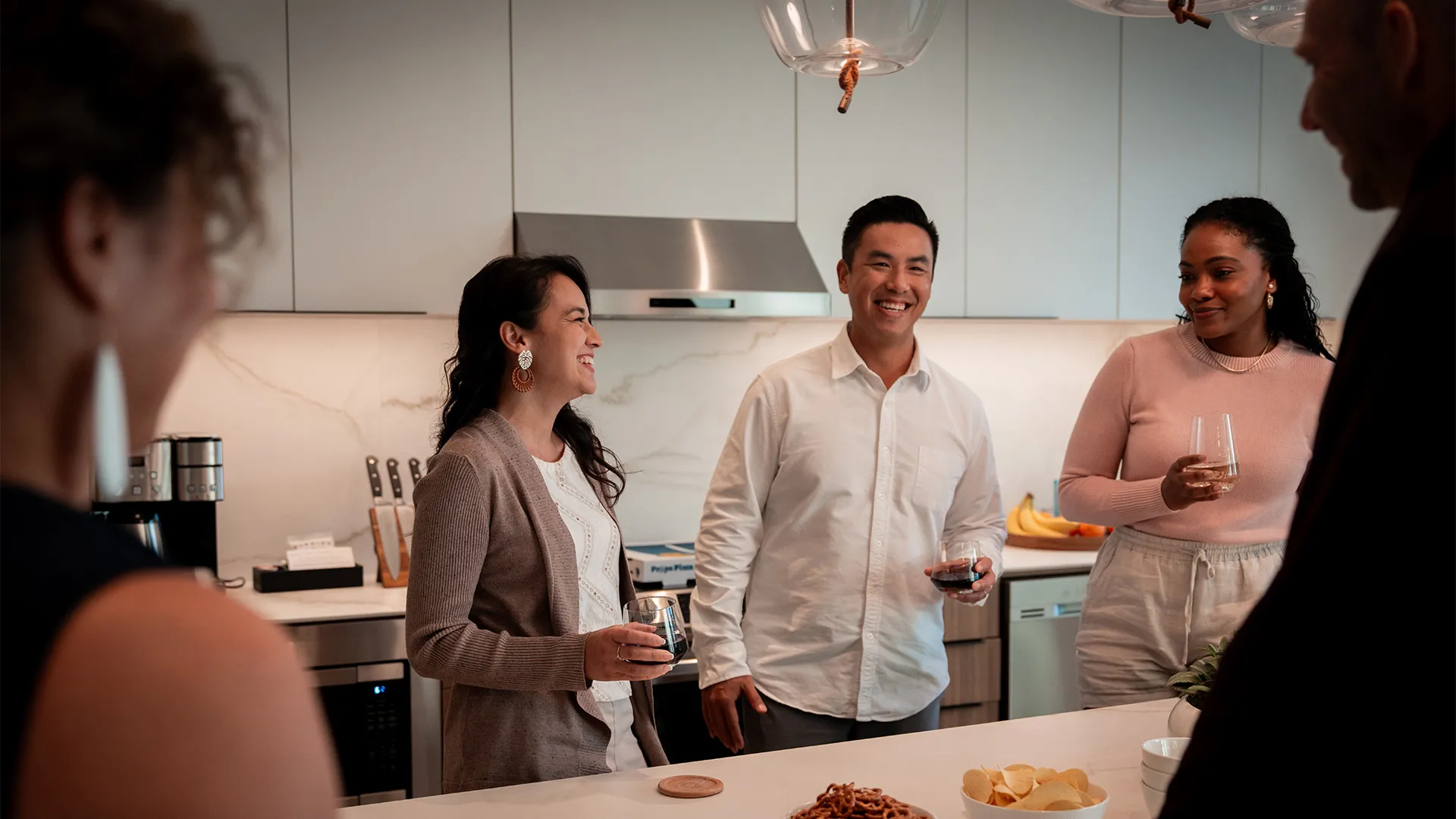 Four people standing in a kitchen chatting and holding drinks with snacks on the counter.