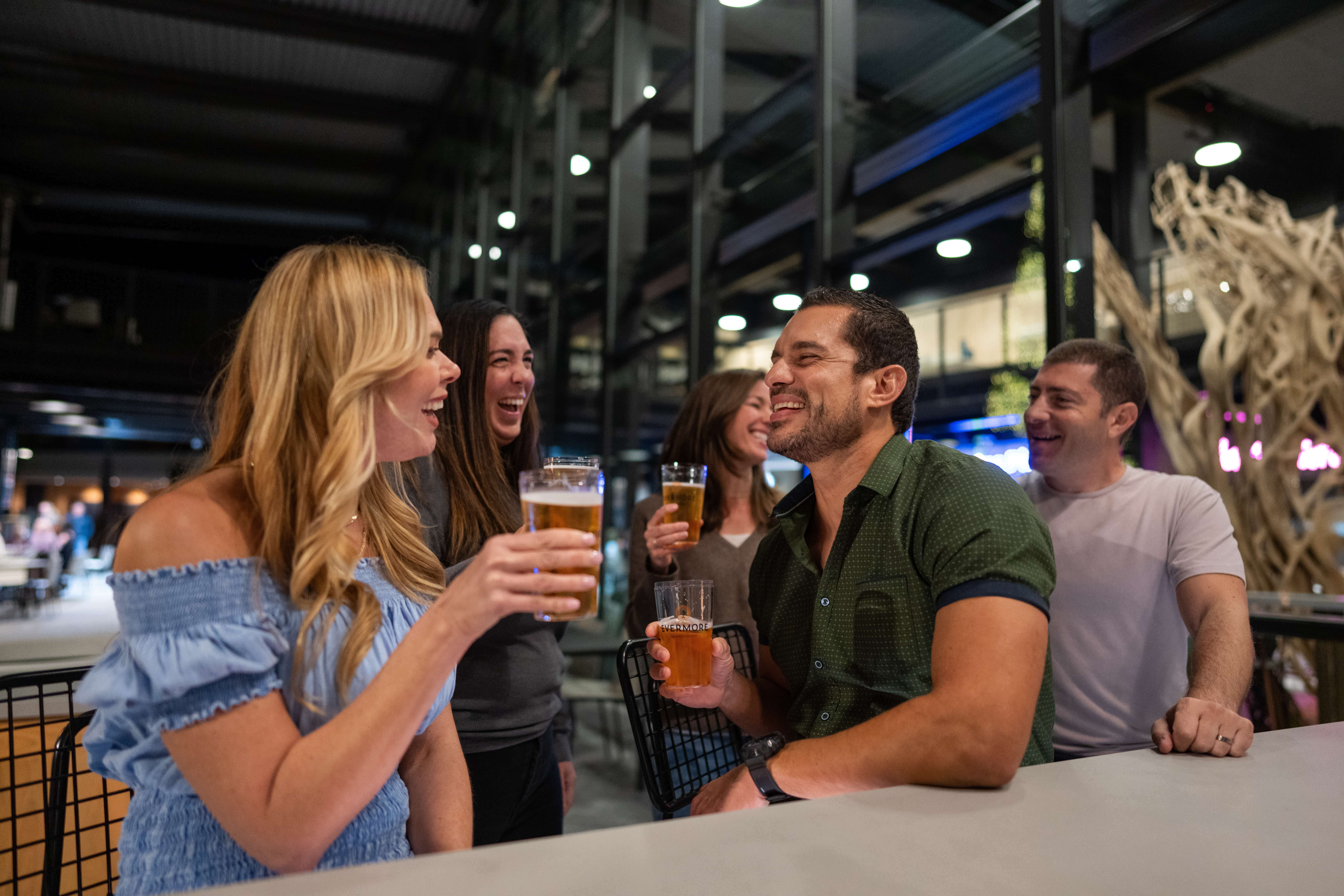 Group of five friends laughing and holding glasses of beer at a bar or restaurant.