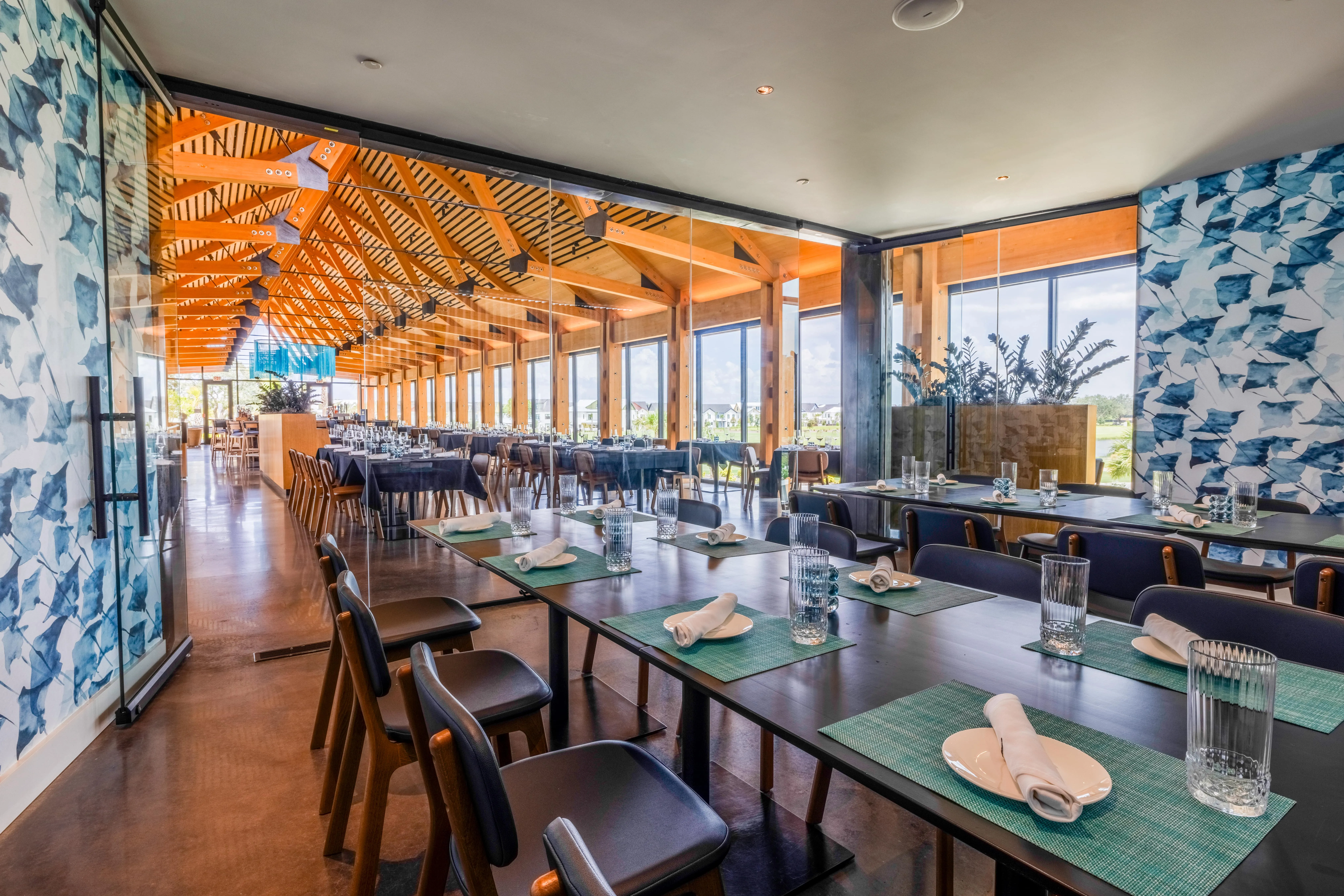 Bright modern restaurant interior with wooden beams, glass walls, and tables set with glasses and napkins.