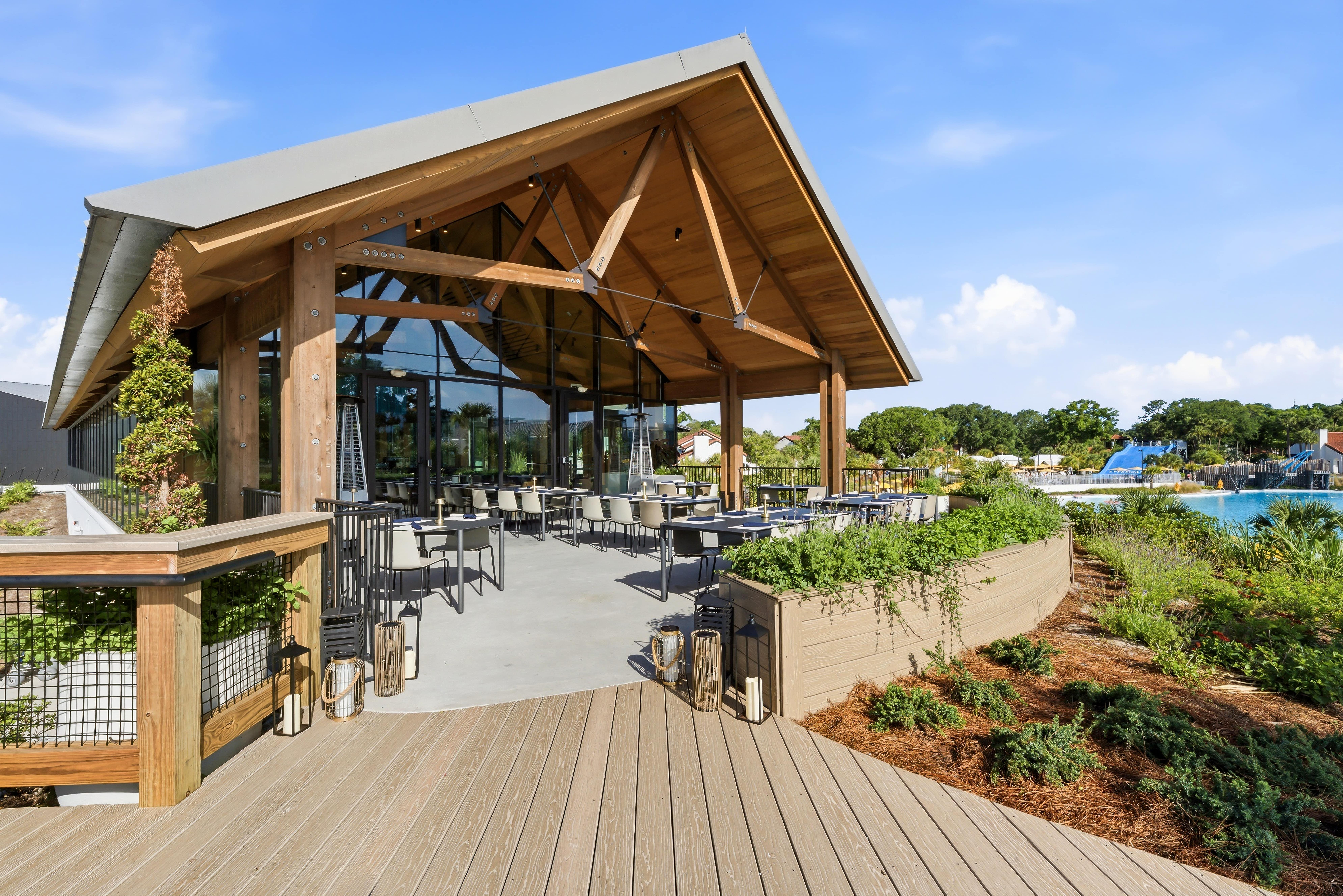 Open-air patio with tables and chairs under a wooden roof next to a landscaped pool area.