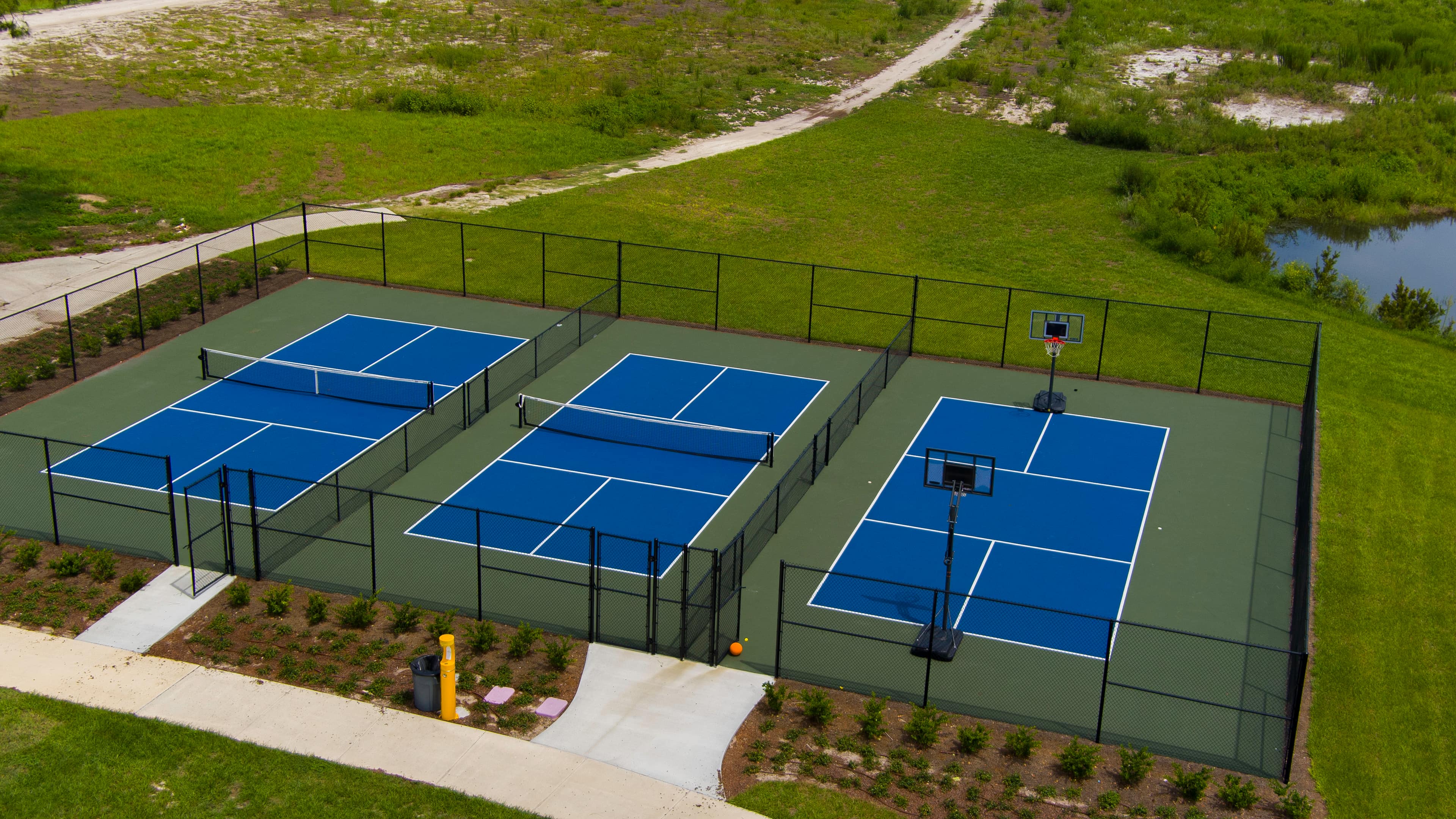 A fenced sports area with two blue tennis courts and one blue basketball court surrounded by grass.