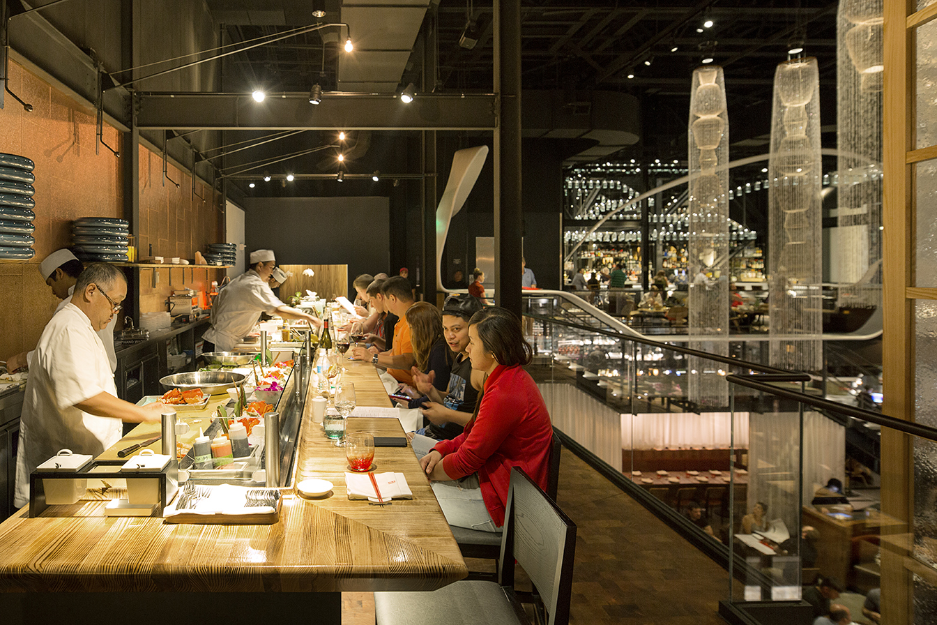 People seated at a sushi bar with chefs preparing food behind the counter in a modern restaurant.