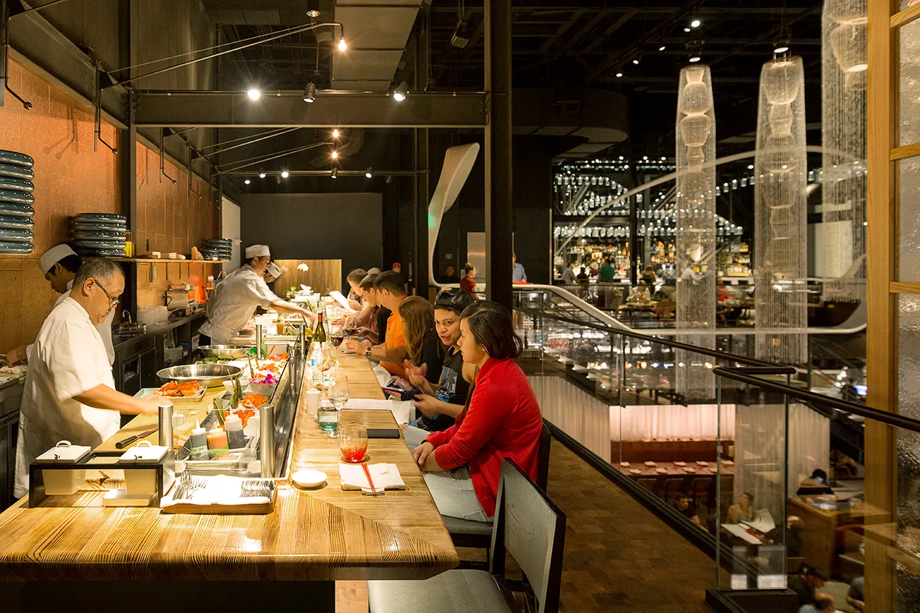 People seated at a sushi bar with chefs preparing food behind the counter in a modern restaurant.