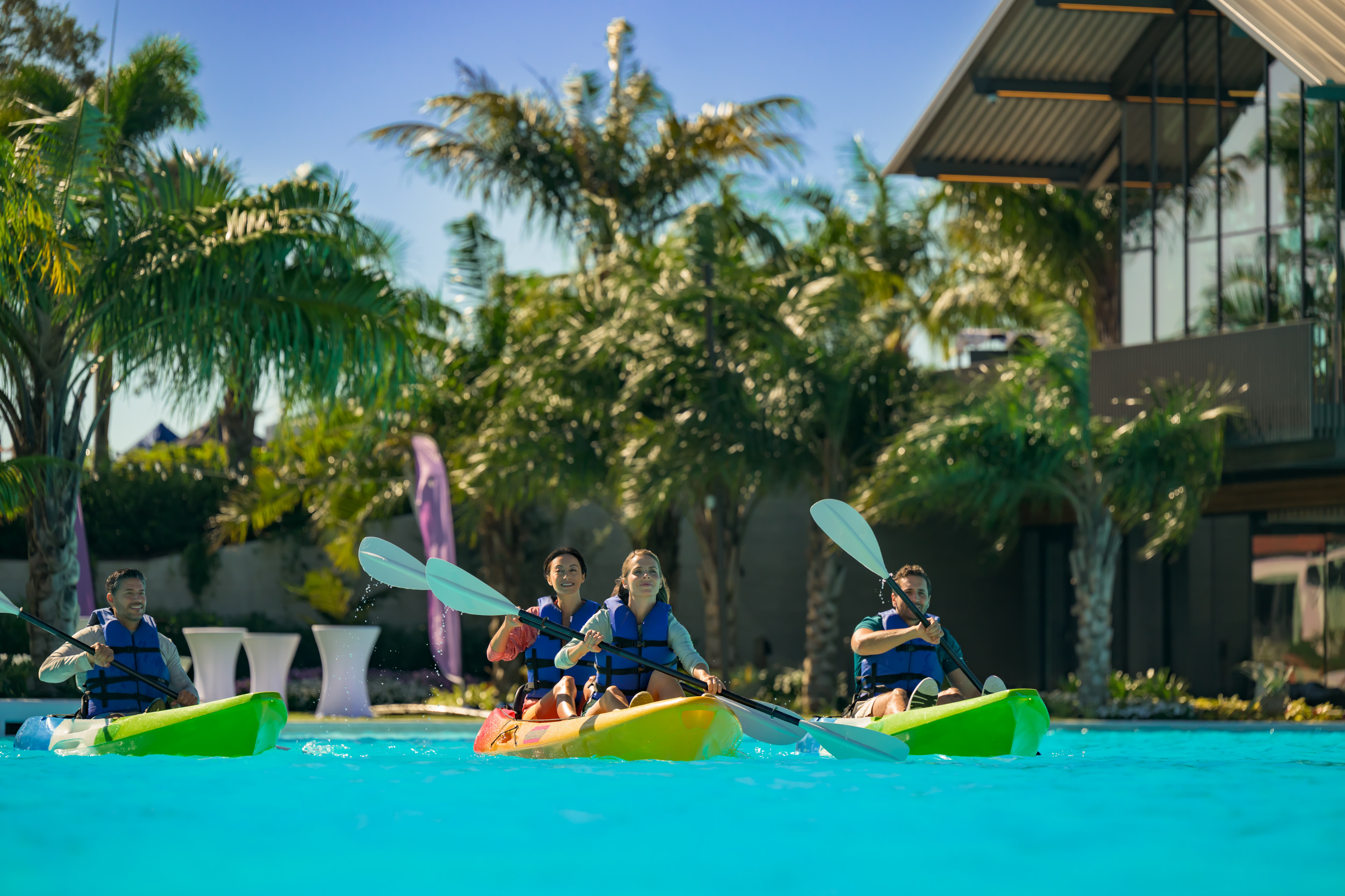 Four people wearing life vests paddling colorful kayaks in a pool with palm trees around.