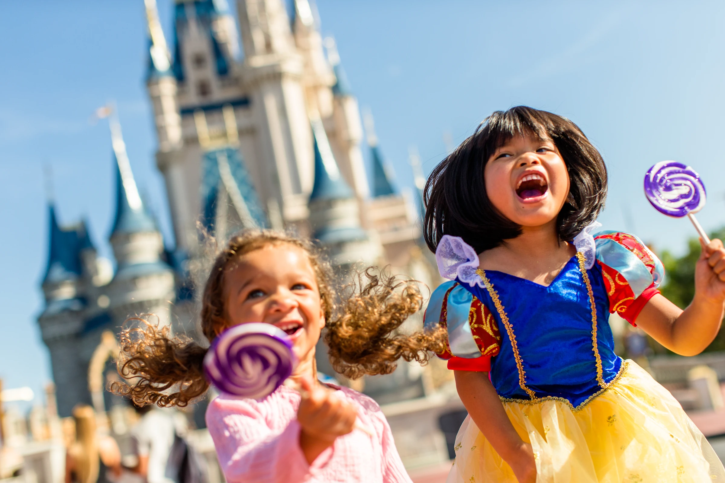 Two joyful girls holding purple swirl lollipops with a fairy tale castle in the background.