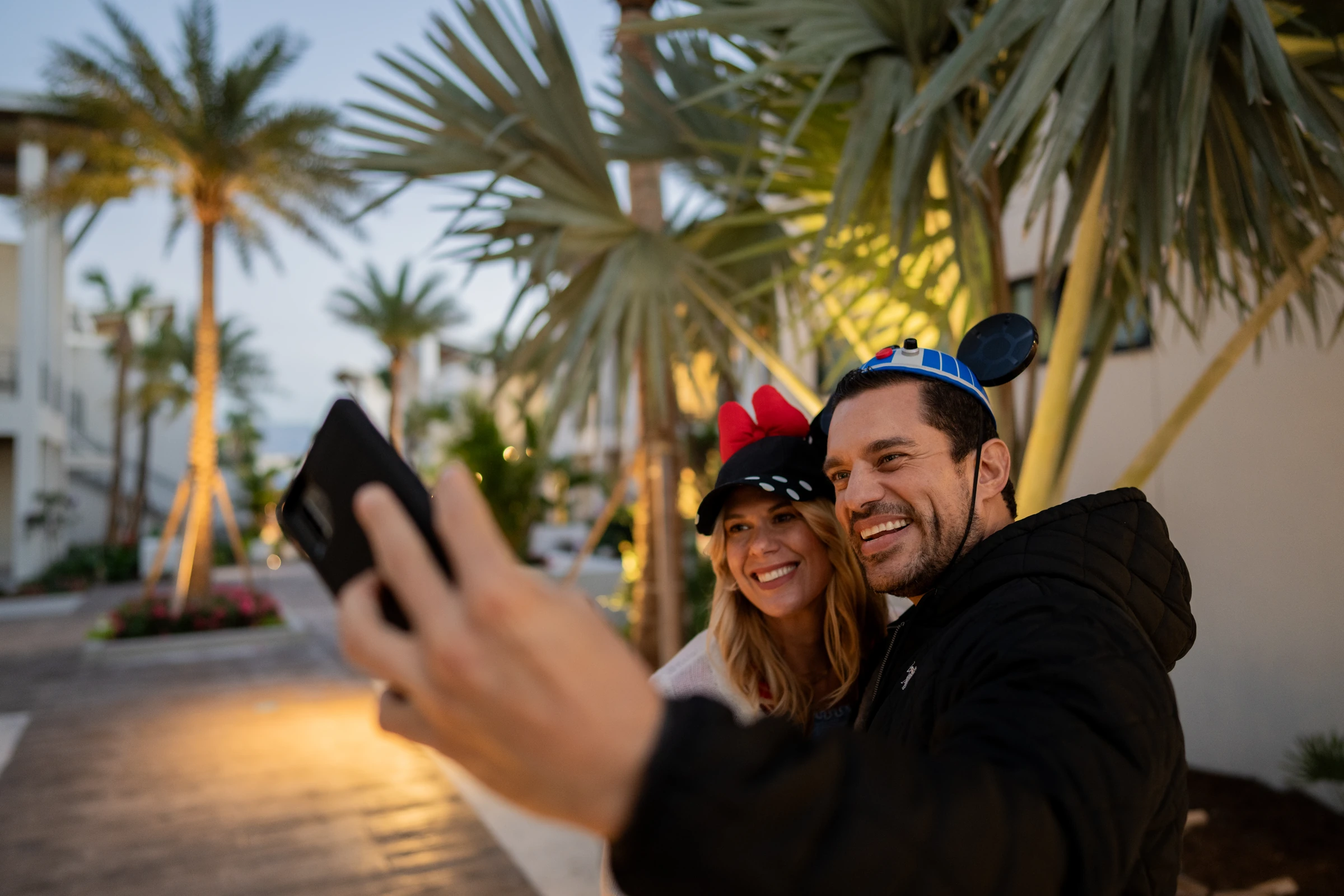 Smiling couple wearing Disney-themed hats takes a selfie outdoors near palm trees at dusk.