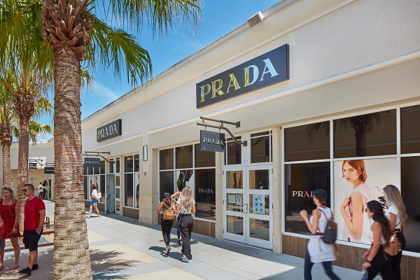 Prada store exterior in sunny outdoor shopping area with people walking by palm trees.