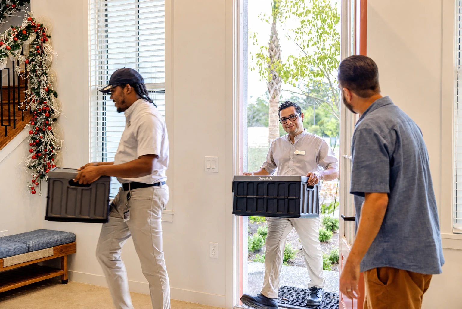 Two delivery men carrying black boxes entering a home with a man greeting them inside.