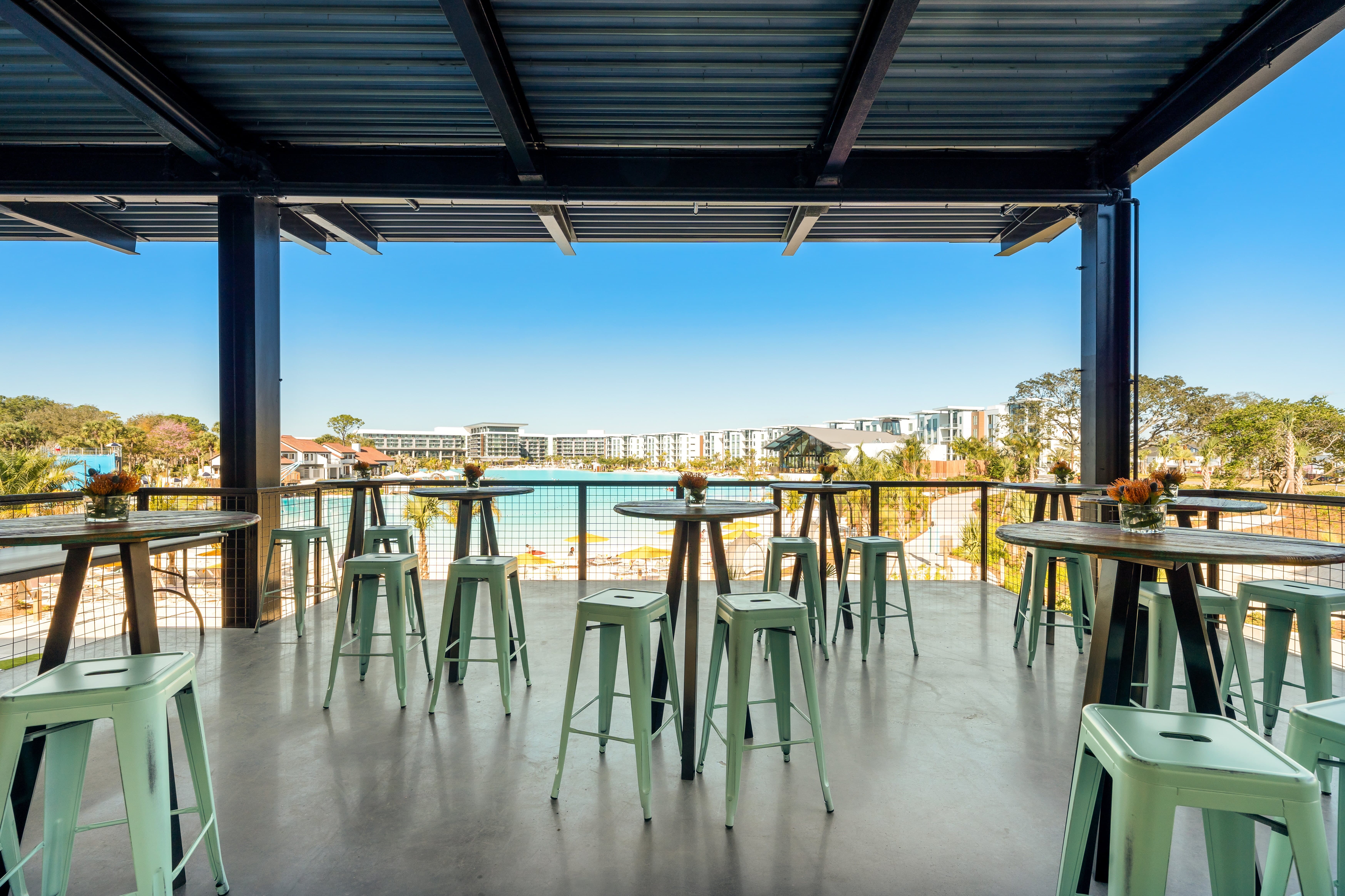 Outdoor covered patio with tables and stools overlooking pool and resort buildings under clear blue sky.