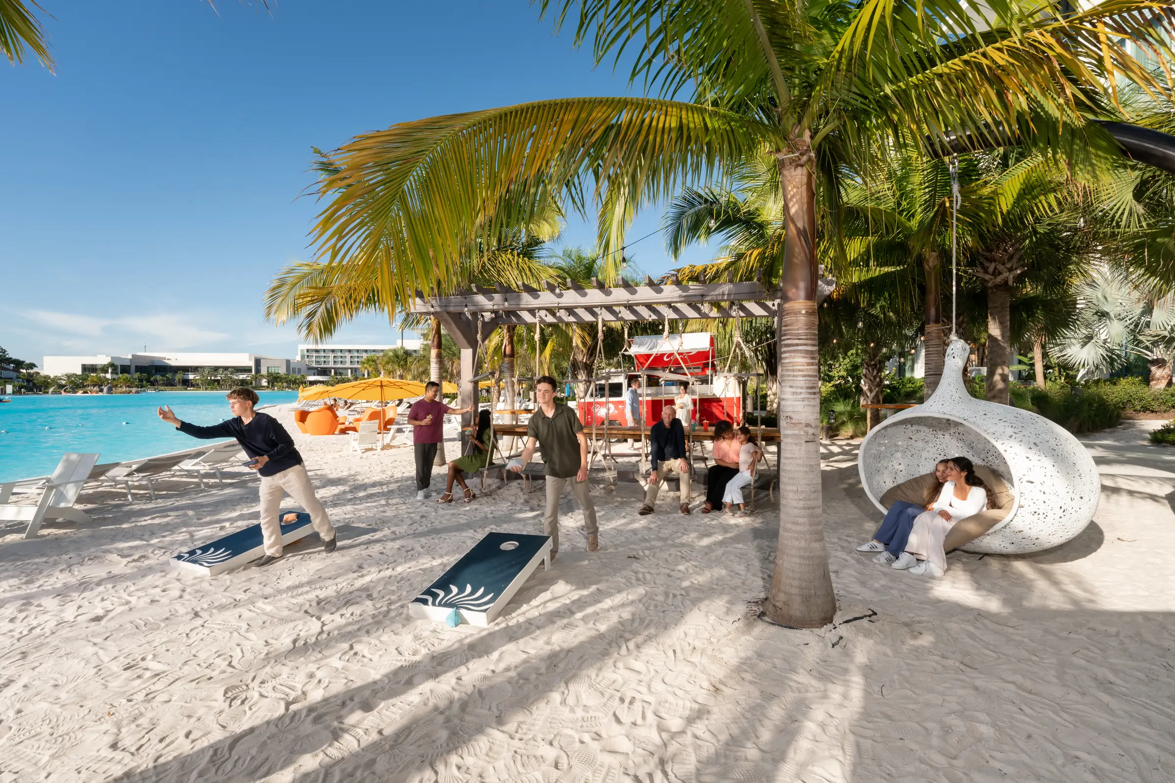 People playing cornhole and relaxing on swings and a pod chair on a sandy beach by a pool.