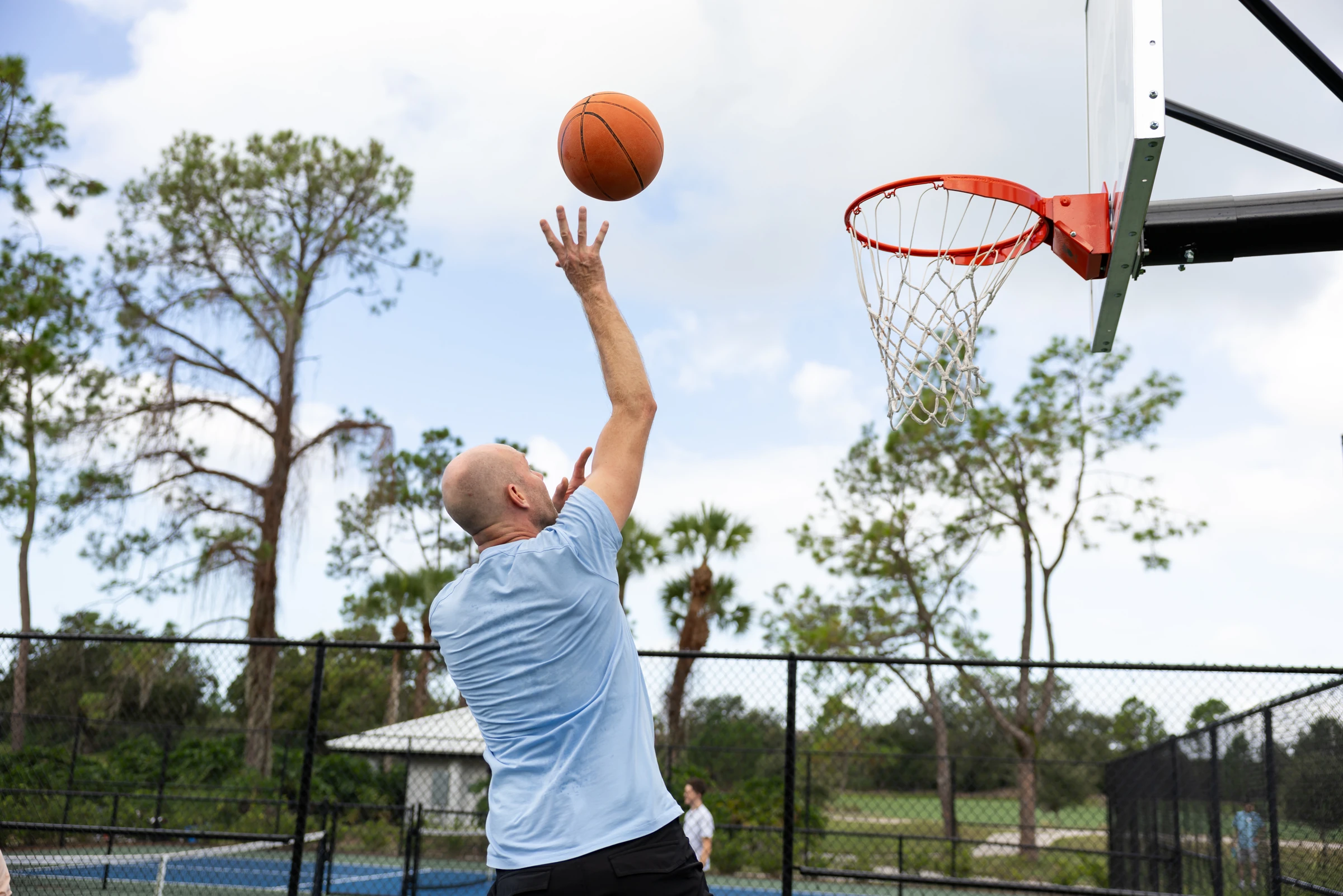 Man in blue shirt shooting a basketball towards the hoop outdoors on a sunny day.