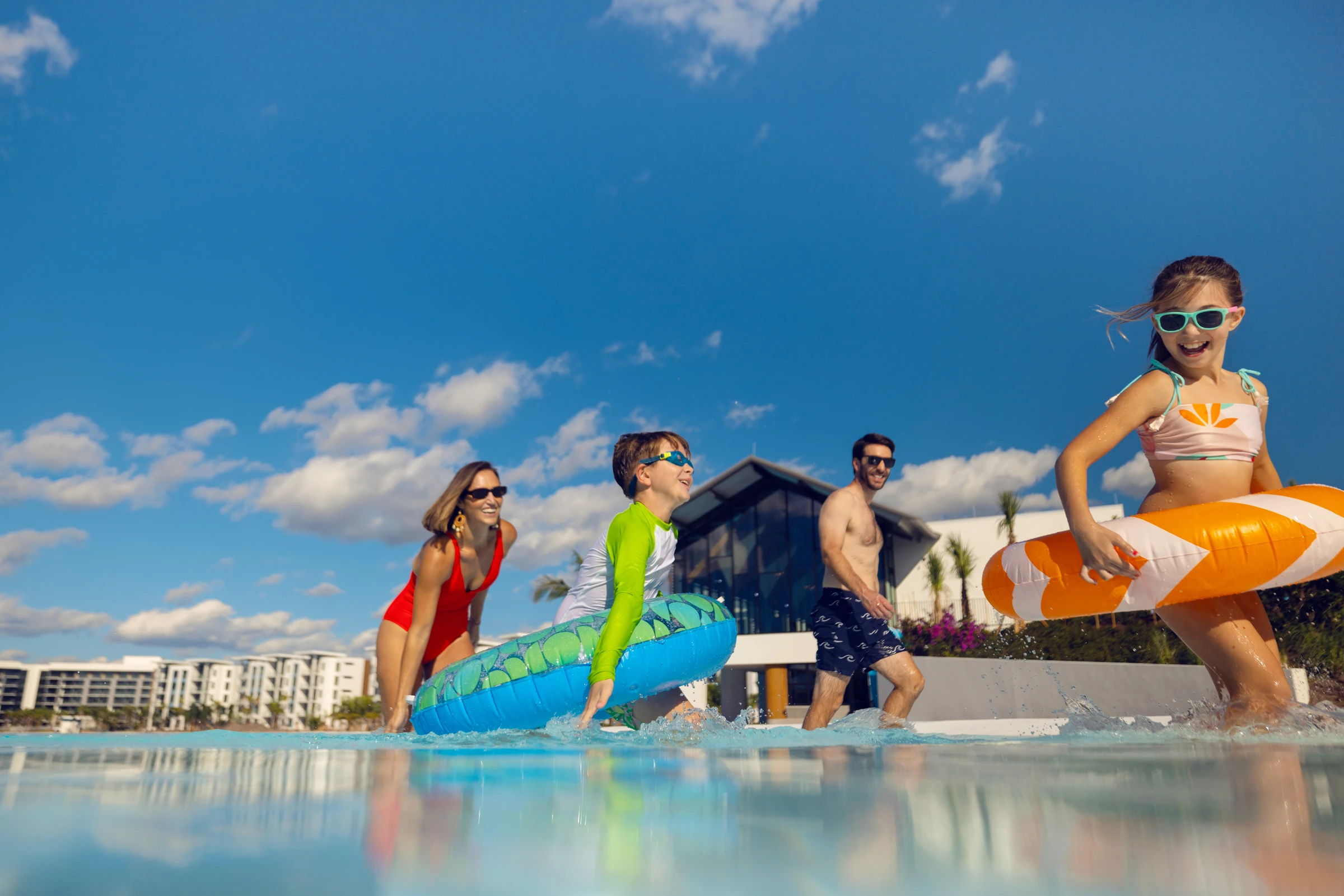 Family of four wearing sunglasses playing in a pool with inflatable swim rings on a sunny day.