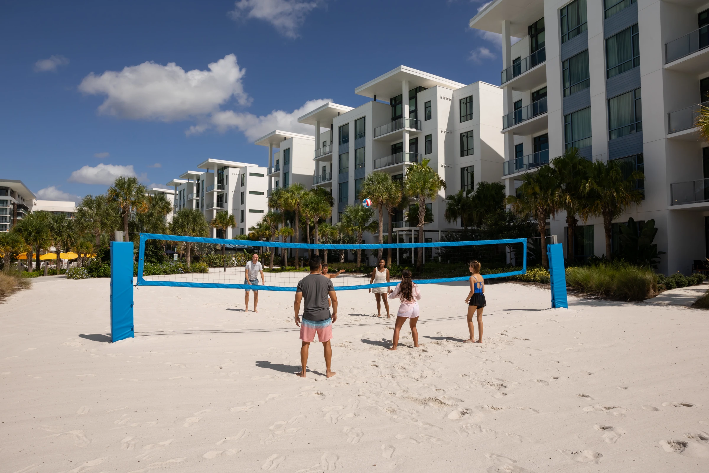 Six people playing beach volleyball with net in front of modern white apartment buildings and palm trees.