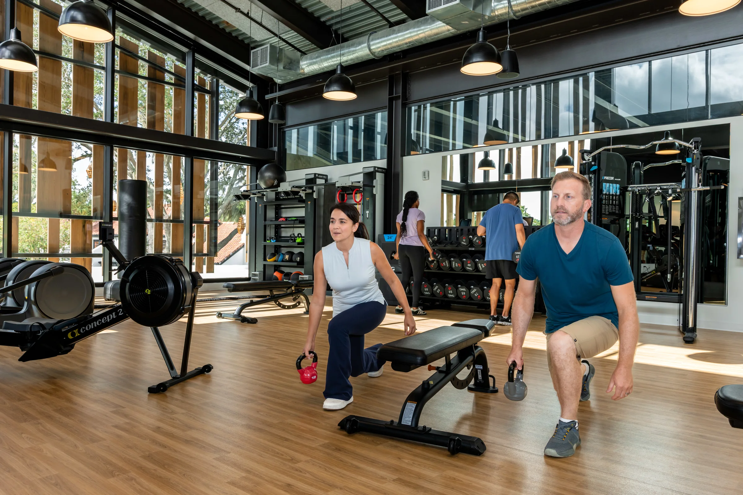 Man and woman doing lunges with kettlebells beside a bench in a bright modern gym.